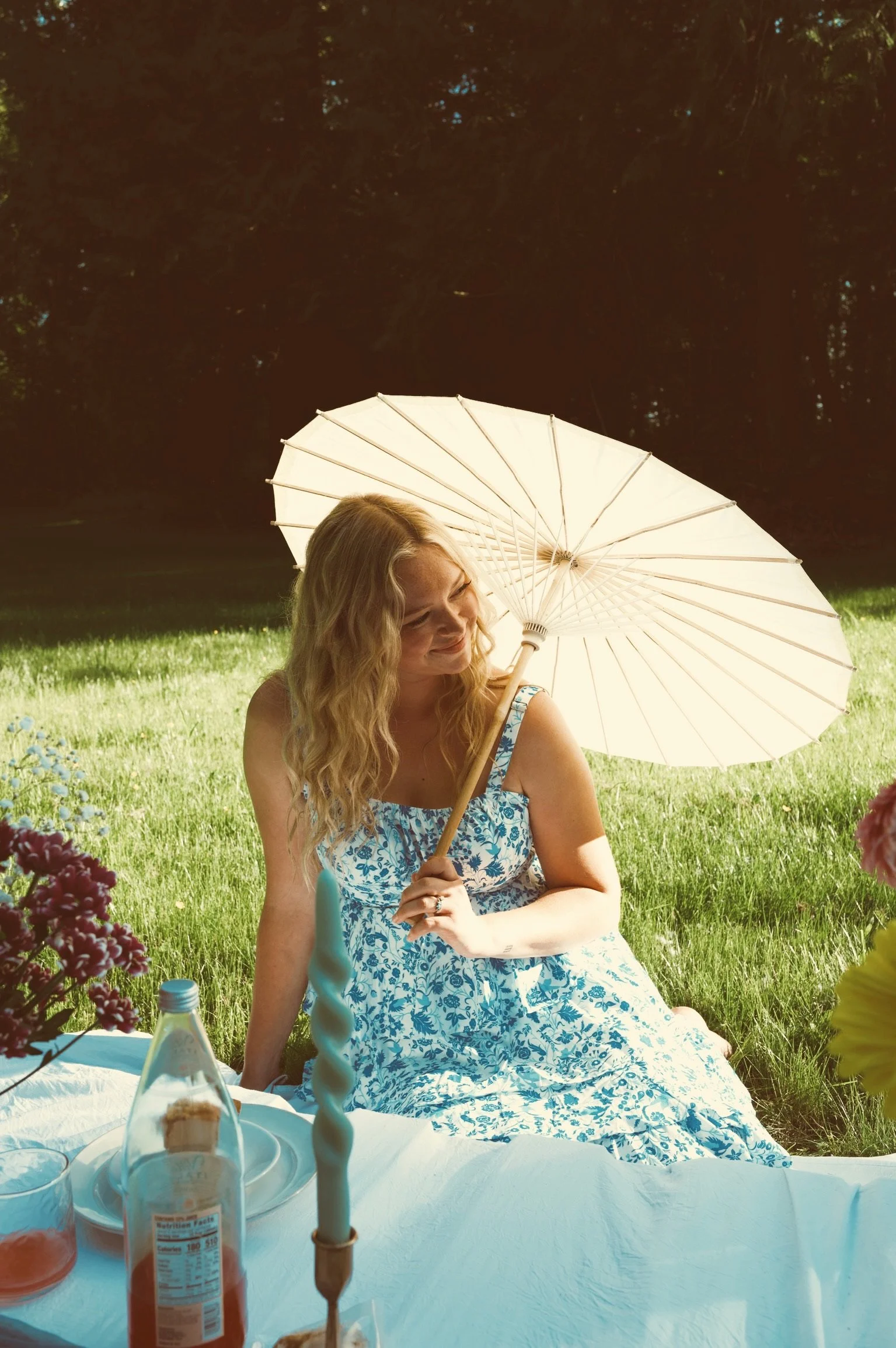 Woman in a blue floral dress sitting beneath a cream parasol in a flower-filled garden.