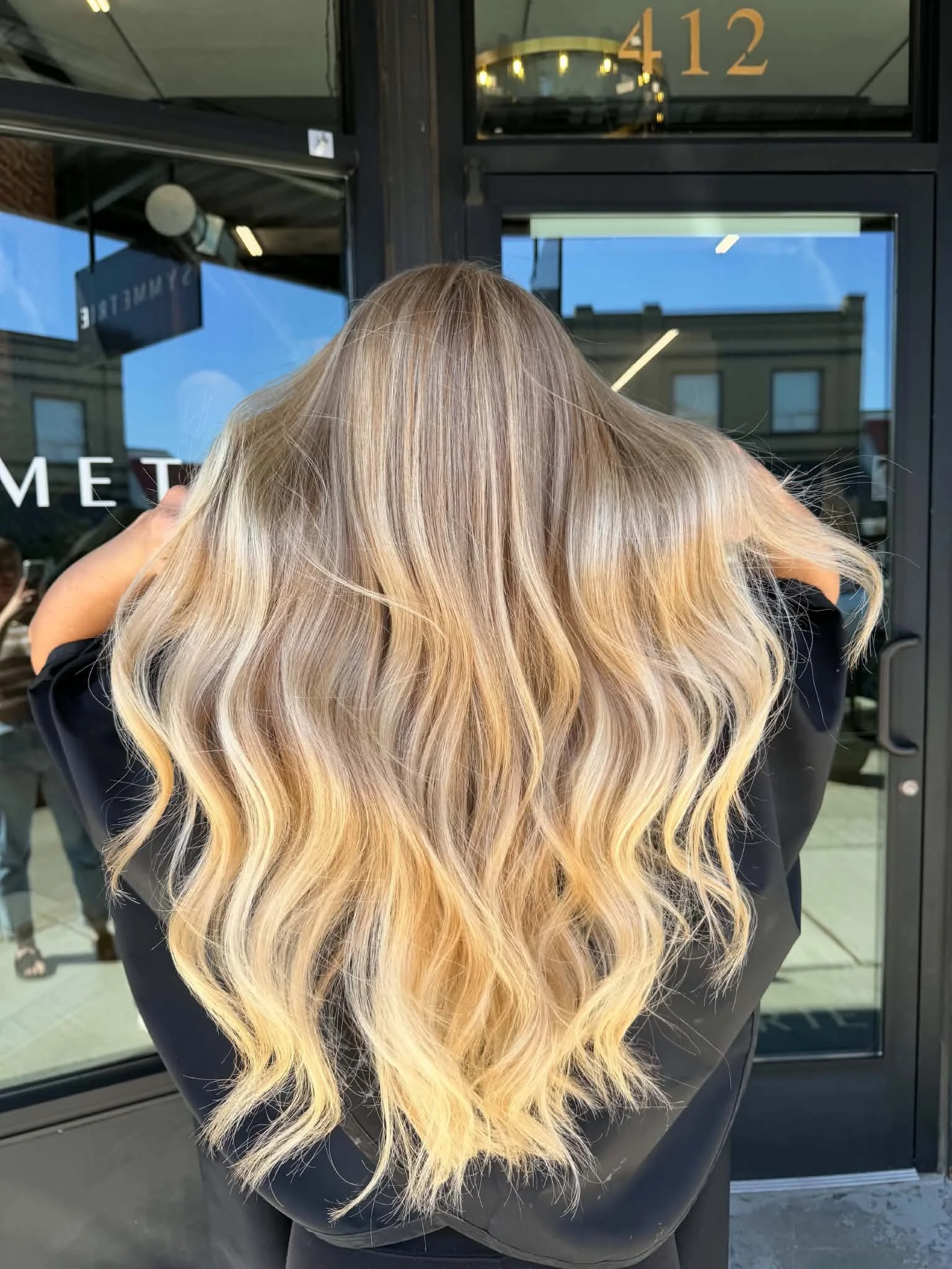 Back view of long blonde balayage with blended roots and soft waves by Caitlyn Deming.