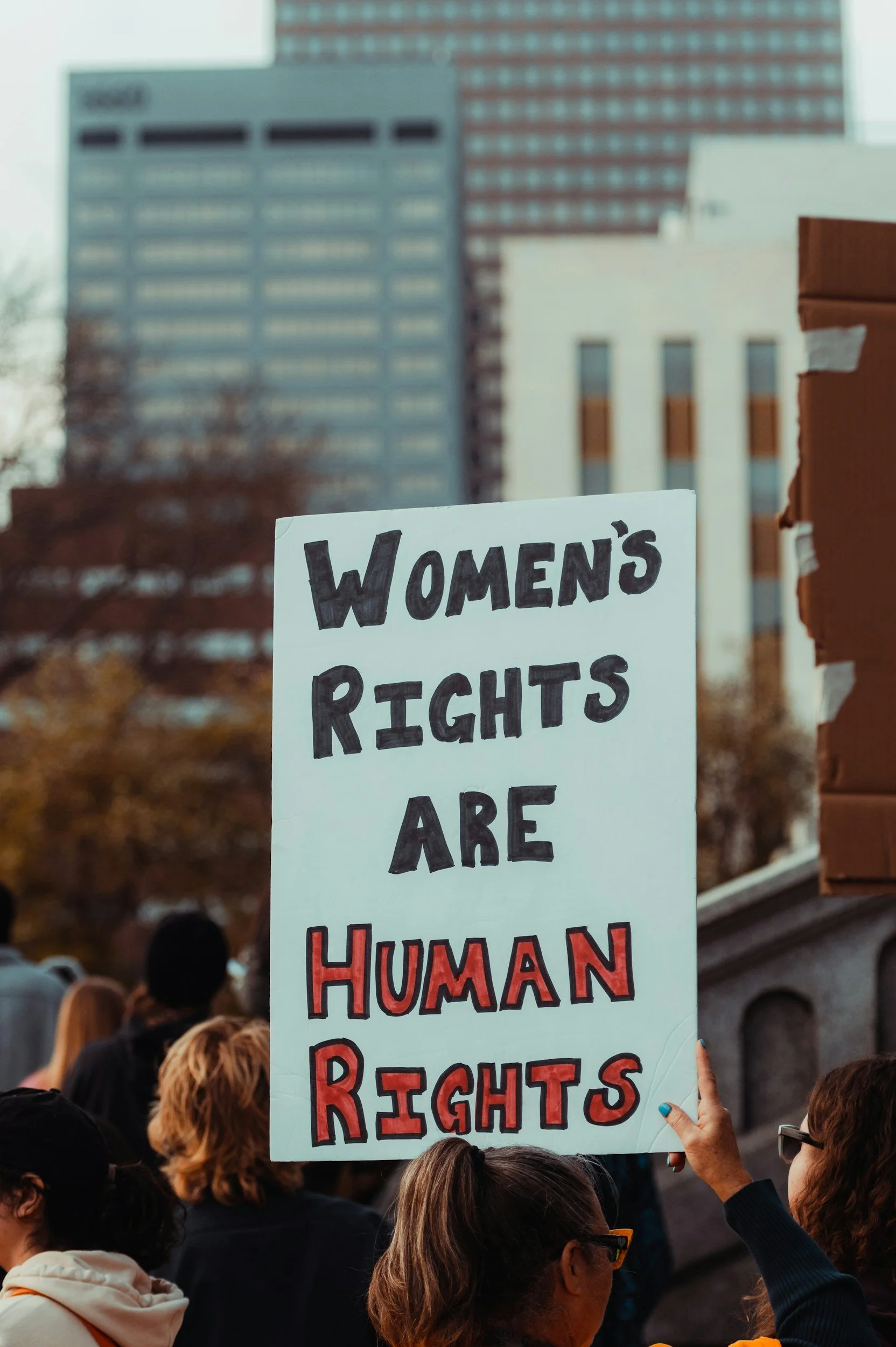 Protestor holding a sign that reads 'Women's Rights Are Human Rights' during a rally with a crowd of people in an urban area with tall buildings in the background.
