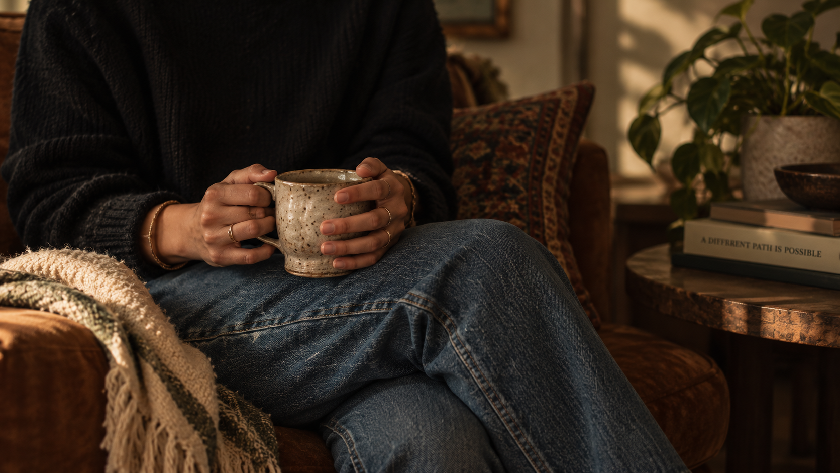 Person sitting on a brown armchair holding a ceramic mug, wearing a black sweater and blue jeans, with a beige knitted blanket on the armrest. In the background, a side table with a plant, some books, and a bowl is visible, in a cozy indoor setting.
