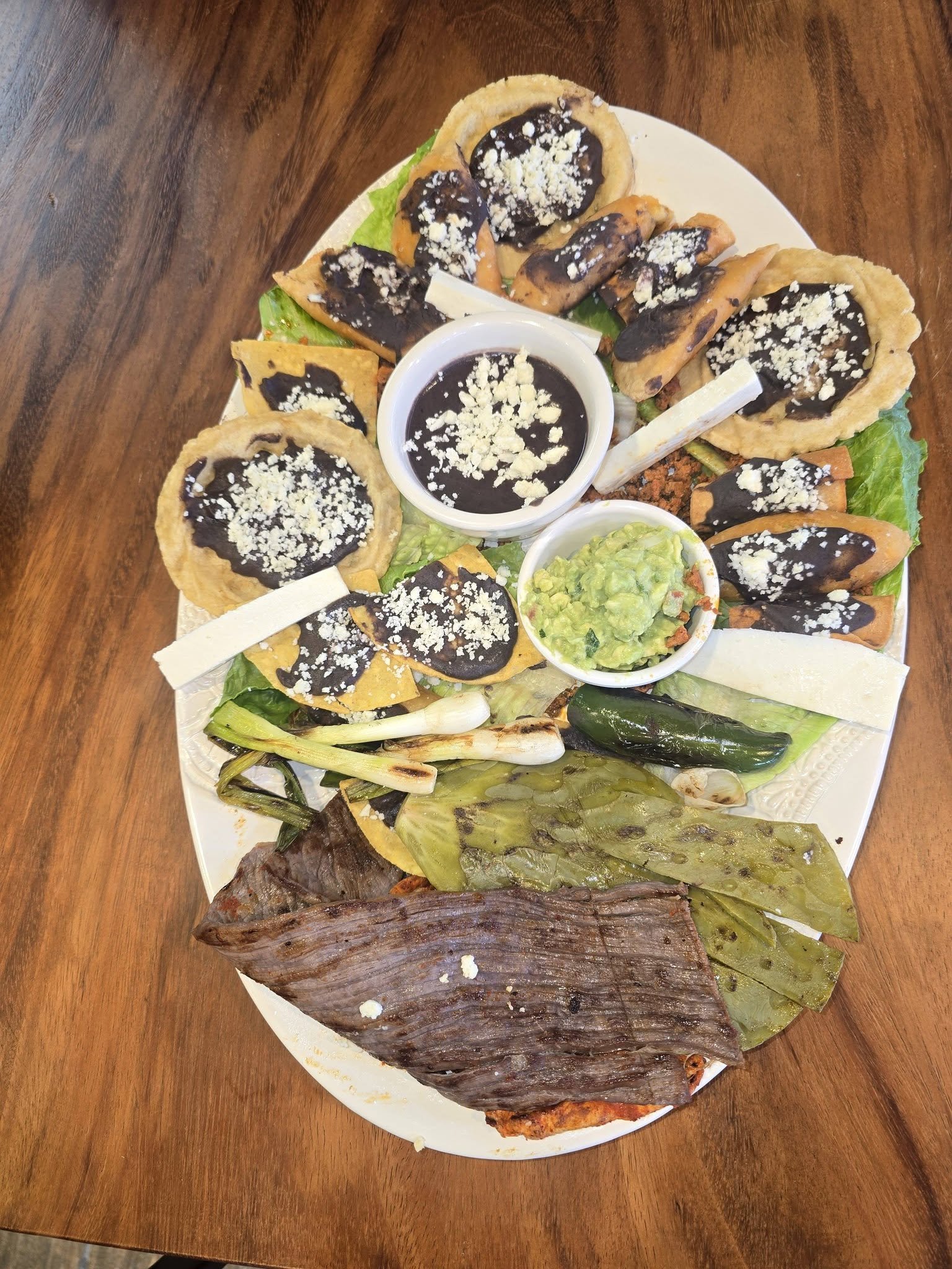 The image shows a large dish of Mexican food features grilled fish topped with banana leaves, accompanied by guacamole, grilled onions, and various tacos with dark sauce and cheese, all served on a white platter on a wooden table.