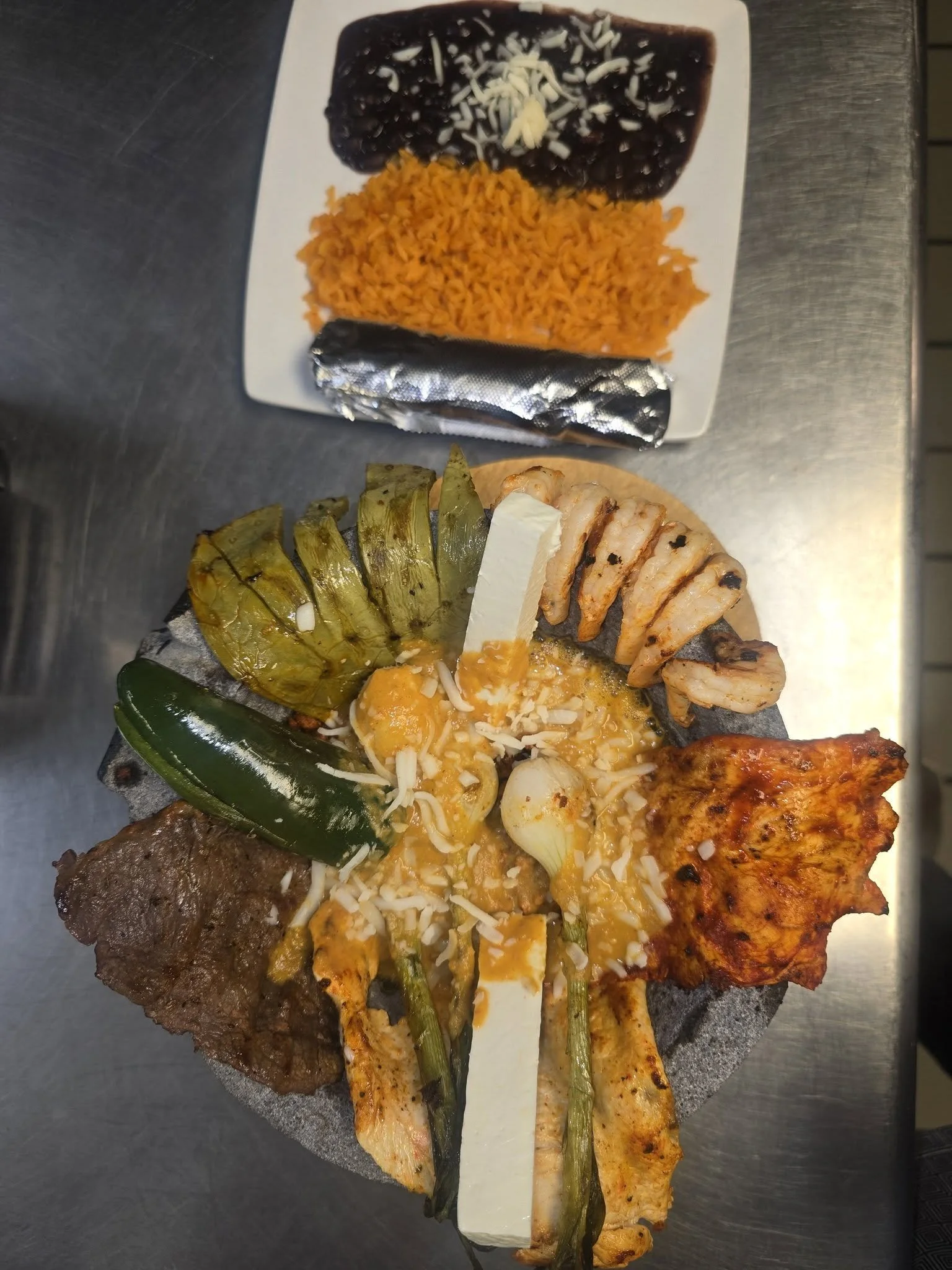 Platter of Mexican dishes including rice, black beans with cheese, grilled chicken with green peppers, beef steak, and grilled fish with a cheese topping, on a dark stone surface.