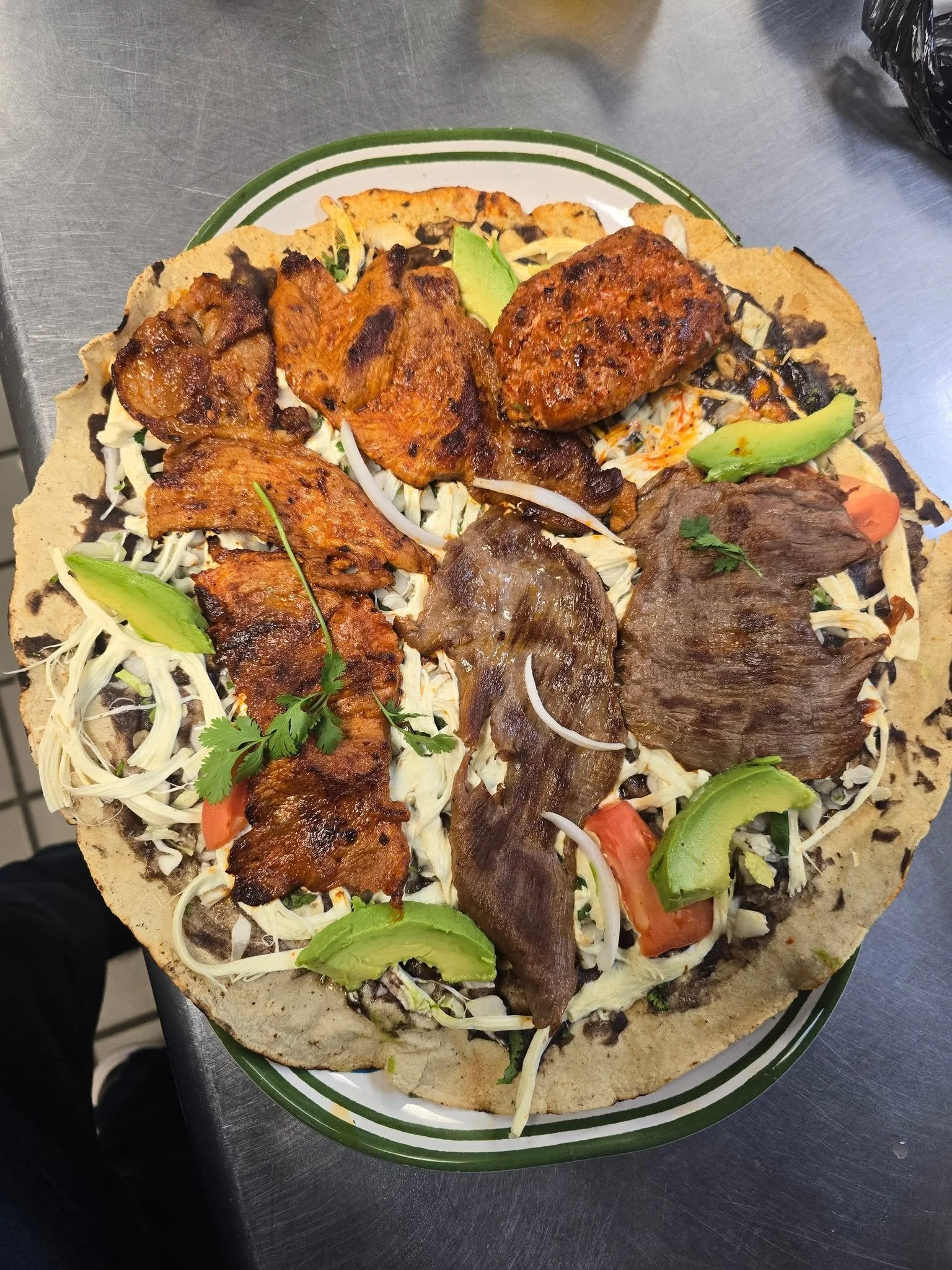 A plate of Mexican food featuring a large, round, thin tortilla topped with shredded cheese, grilled meat, chicken, beef, and vegetables such as avocado slices, tomato, onion, and cilantro.
