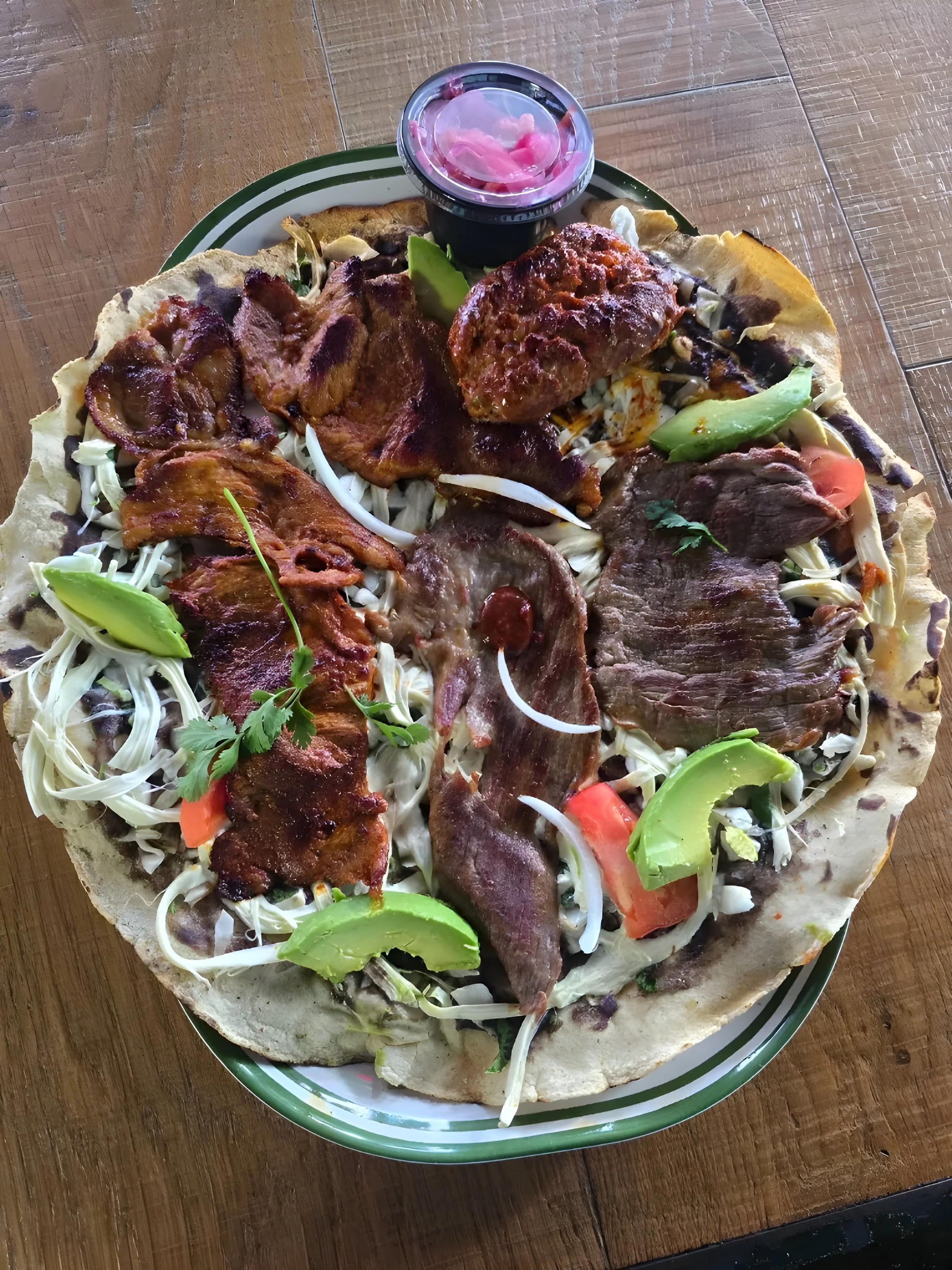 A large pizza topped with various meats, including sliced beef, chicken, and pork, along with vegetables such as avocado, tomato, and bean sprouts, served on a green and white plate on a wooden table. There is a small container of pickled pink onions