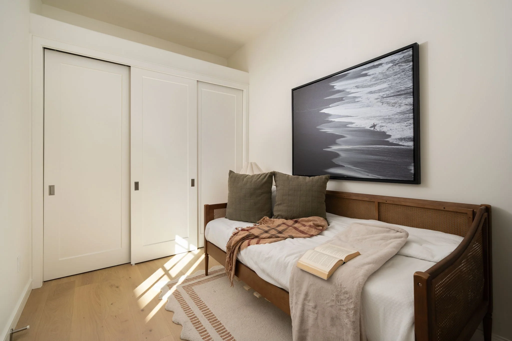 A bedroom with white walls, a wooden bed with a checked pillow and blanket, an open book, a beige carpet, and a large framed black and white beach scene photograph on the wall. There is a closet with white sliding doors.