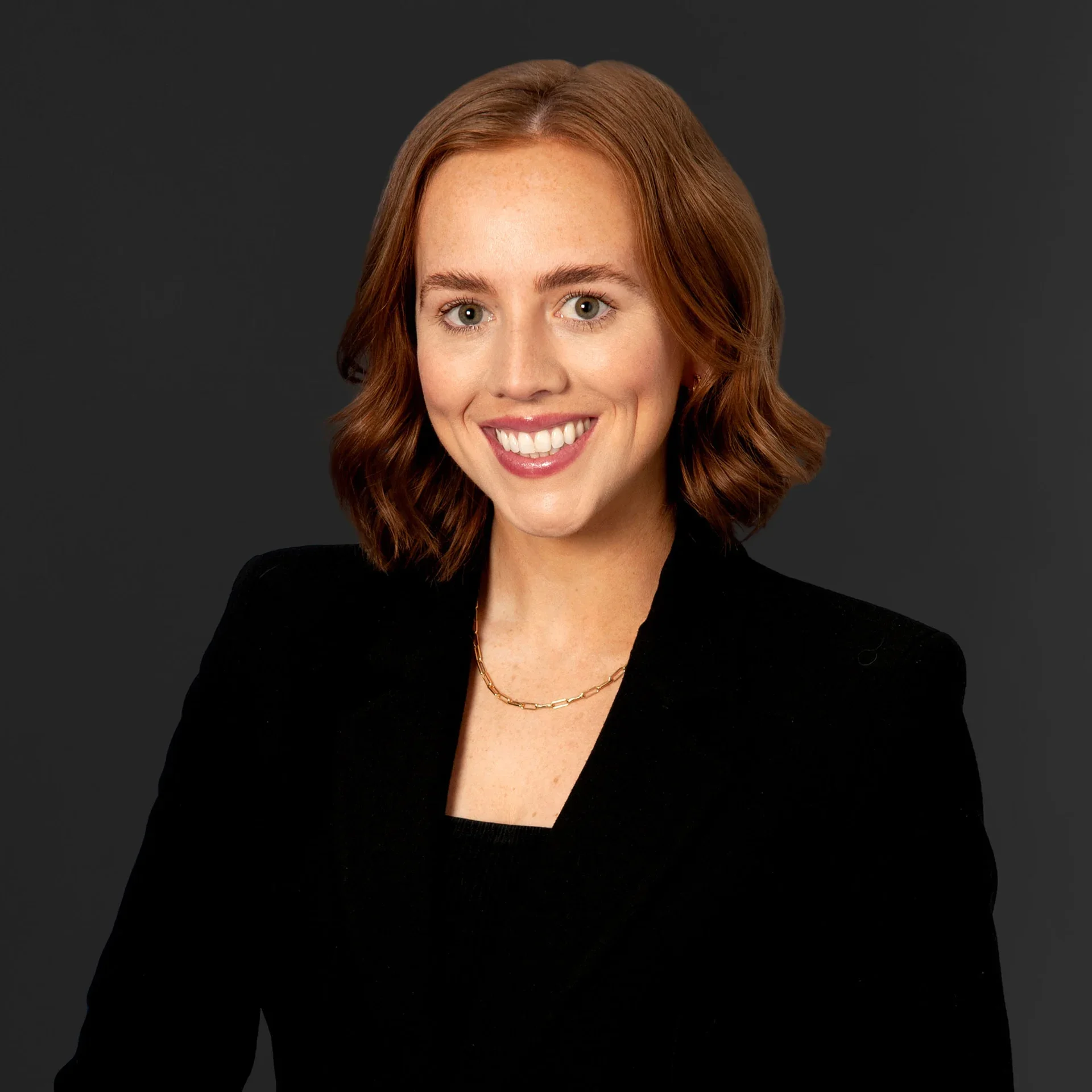A young woman with shoulder-length red hair, wearing a black blazer and a gold necklace, smiling against a dark gray background.
