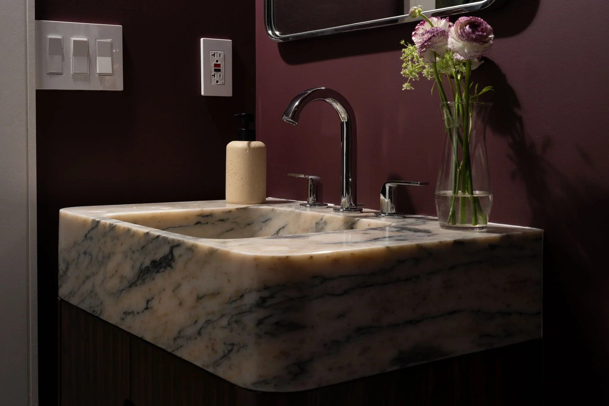 A modern bathroom sink with a marble countertop, a chrome faucet, a beige soap dispenser, and a glass vase with pink and purple flowers against a dark maroon wall.