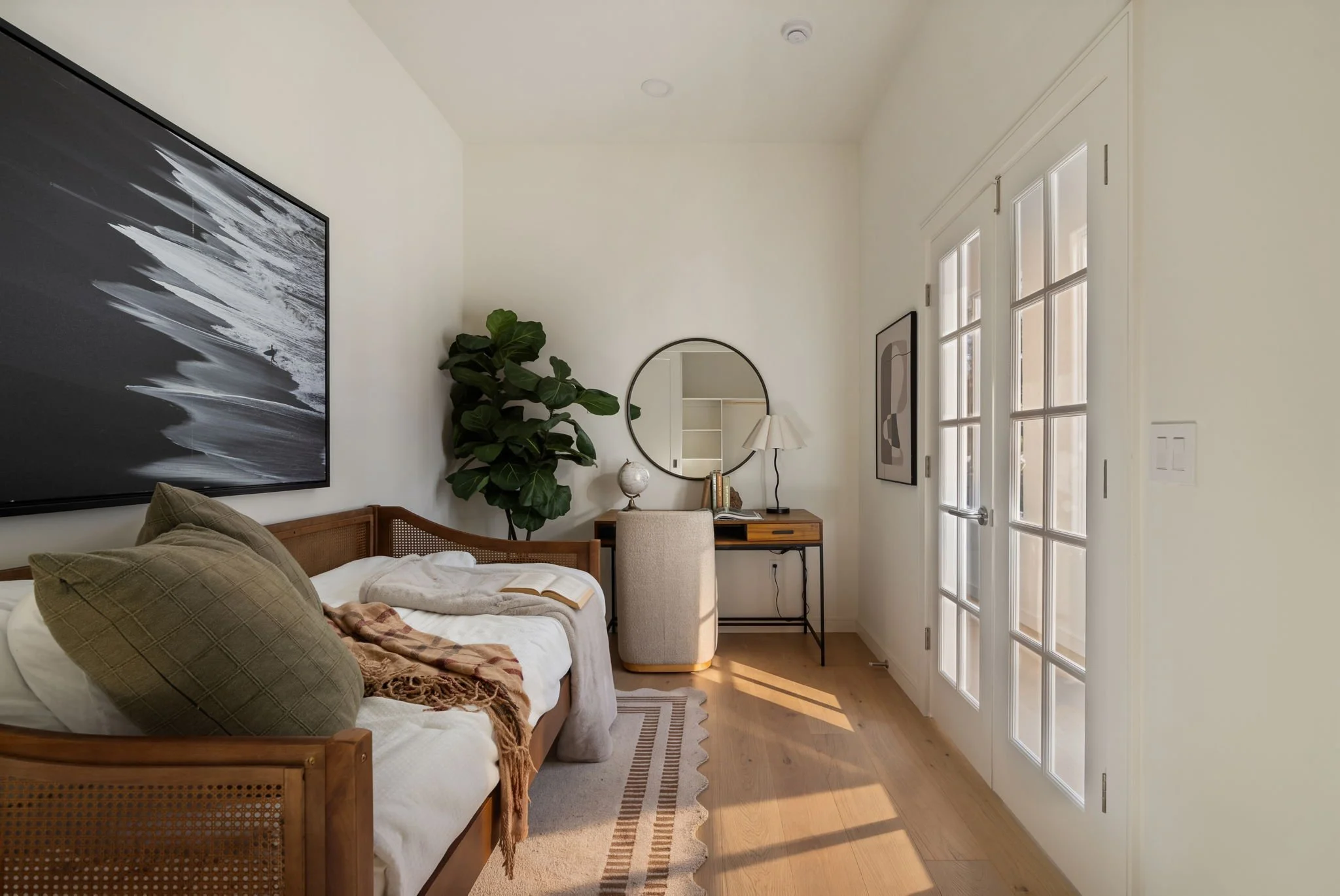 A cozy bedroom with a bed, large framed black and white artwork, a potted plant, a round mirror, a desk with books, a table lamp, and a beige chair in front of a windowed door.