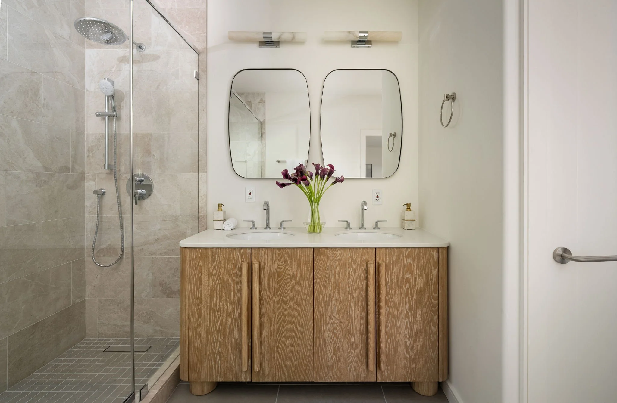 Bathroom with a shower area on the left and a double sink vanity with mirrors and a vase of purple flowers on the right.