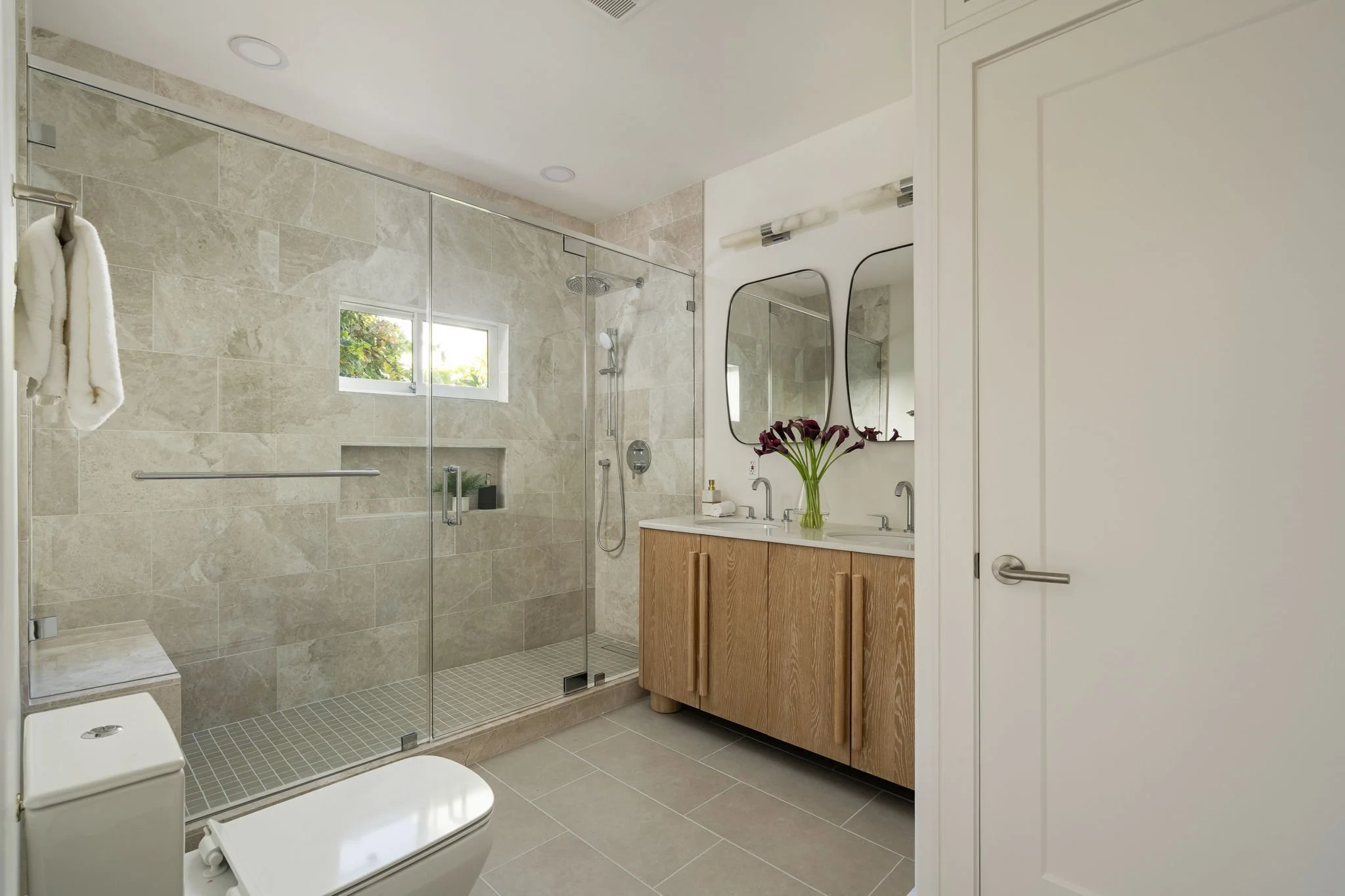 Modern bathroom with a walk-in glass shower, a wooden vanity with a white sink, a countertop mirror, a vase of purple calla lilies, a white toilet, and a small window with greenery outside.