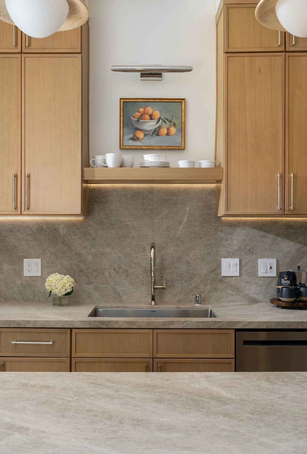 A modern kitchen with wooden cabinets, a granite countertop, a stainless steel sink, and a backsplash with electrical outlets. Decorative items include a small potted flower arrangement, a bowl of oranges, and white cups and dishes on a floating shelf above the sink. A painting of oranges in a bowl hangs on the wall above the shelf.