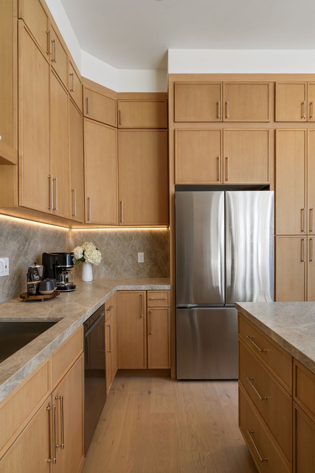 Modern kitchen with wooden cabinets, stainless steel refrigerator, beige countertops, and a coffee station with a mug, coffee maker, and a vase of white flowers.