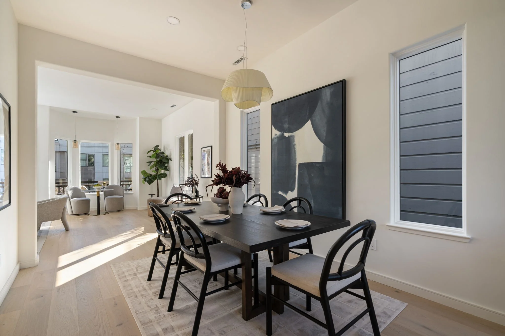Modern dining room with black dining table set for six, surrounded by black chairs with light-colored cushions. A large abstract black and white artwork decorates the white wall. There are windows with gray shutters, a hanging pendant light, and a po