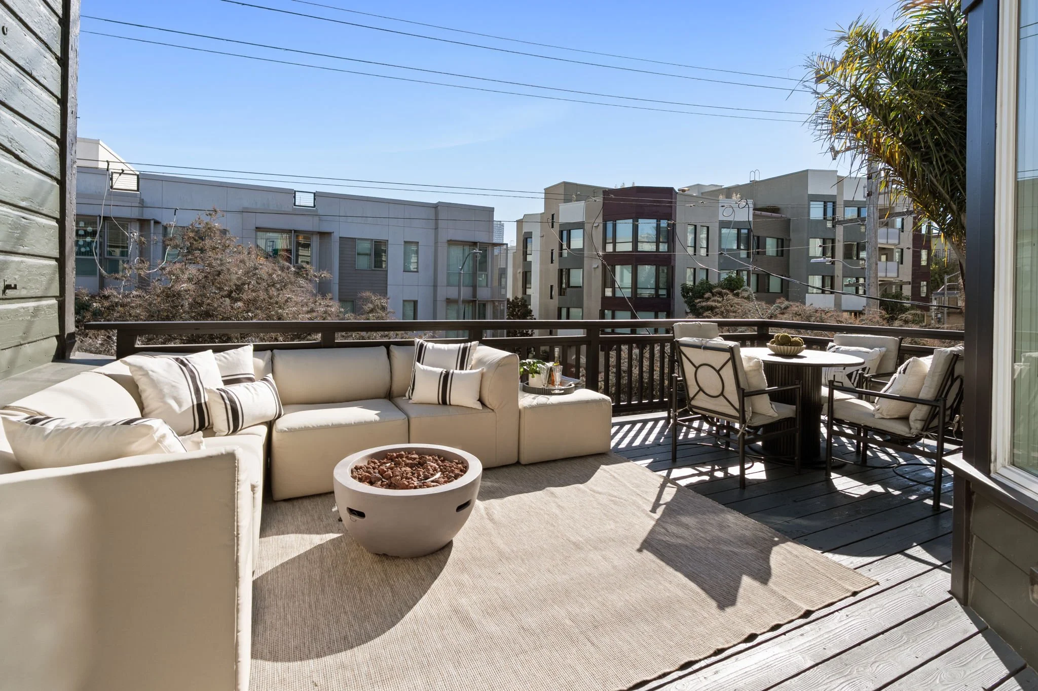 View of a balcony with outdoor seating, including a sectional sofa with pillows, a round table with chairs, and nearby plants, overlooking neighboring modern multi-story residential buildings, under a clear blue sky.