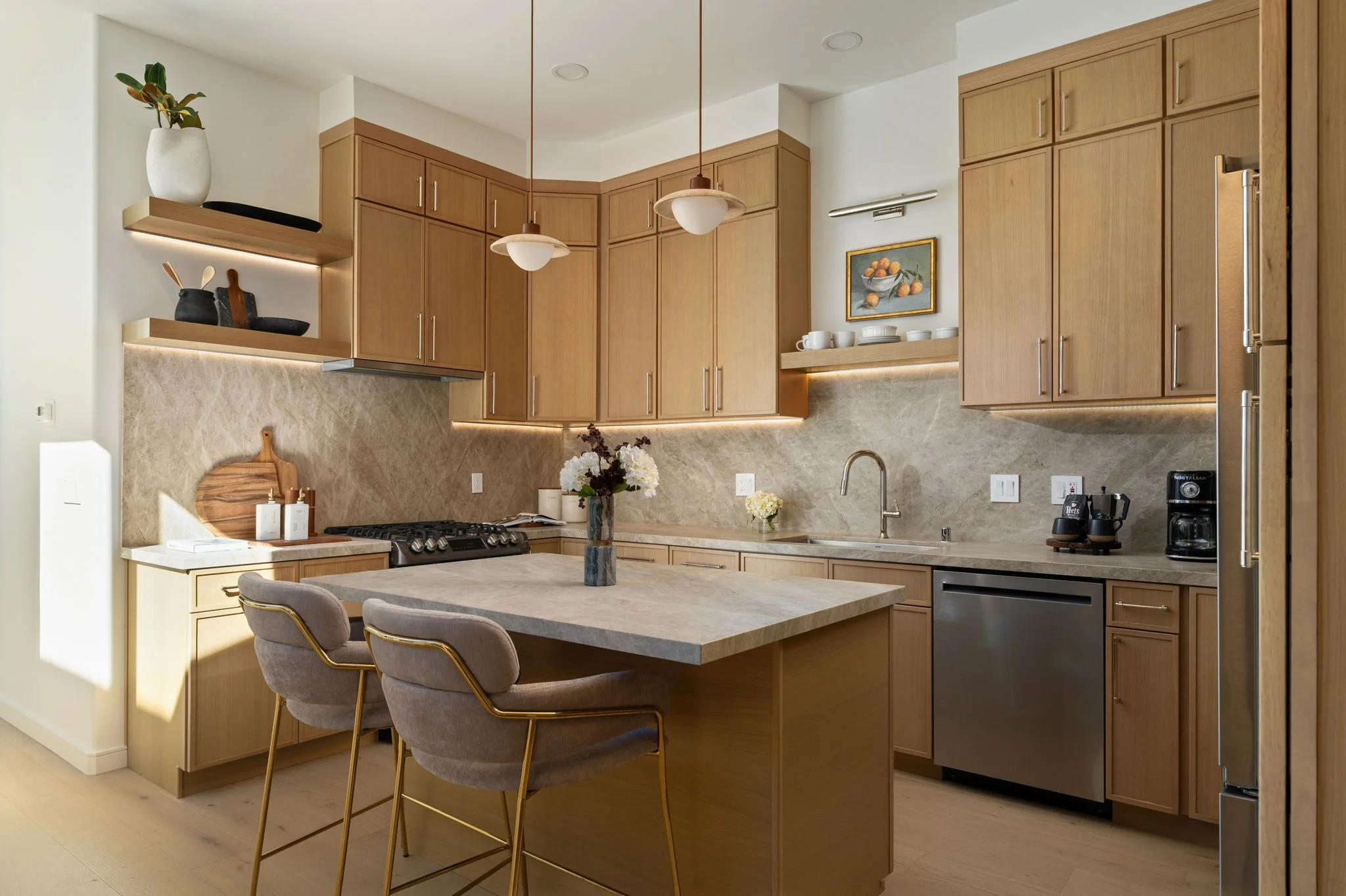 Modern kitchen with wooden cabinets, beige countertops, and two pendant lights hanging above a kitchen island with a bouquet of white flowers. There are open shelves with decor, a built-in oven, and a stainless steel refrigerator. The kitchen has a l