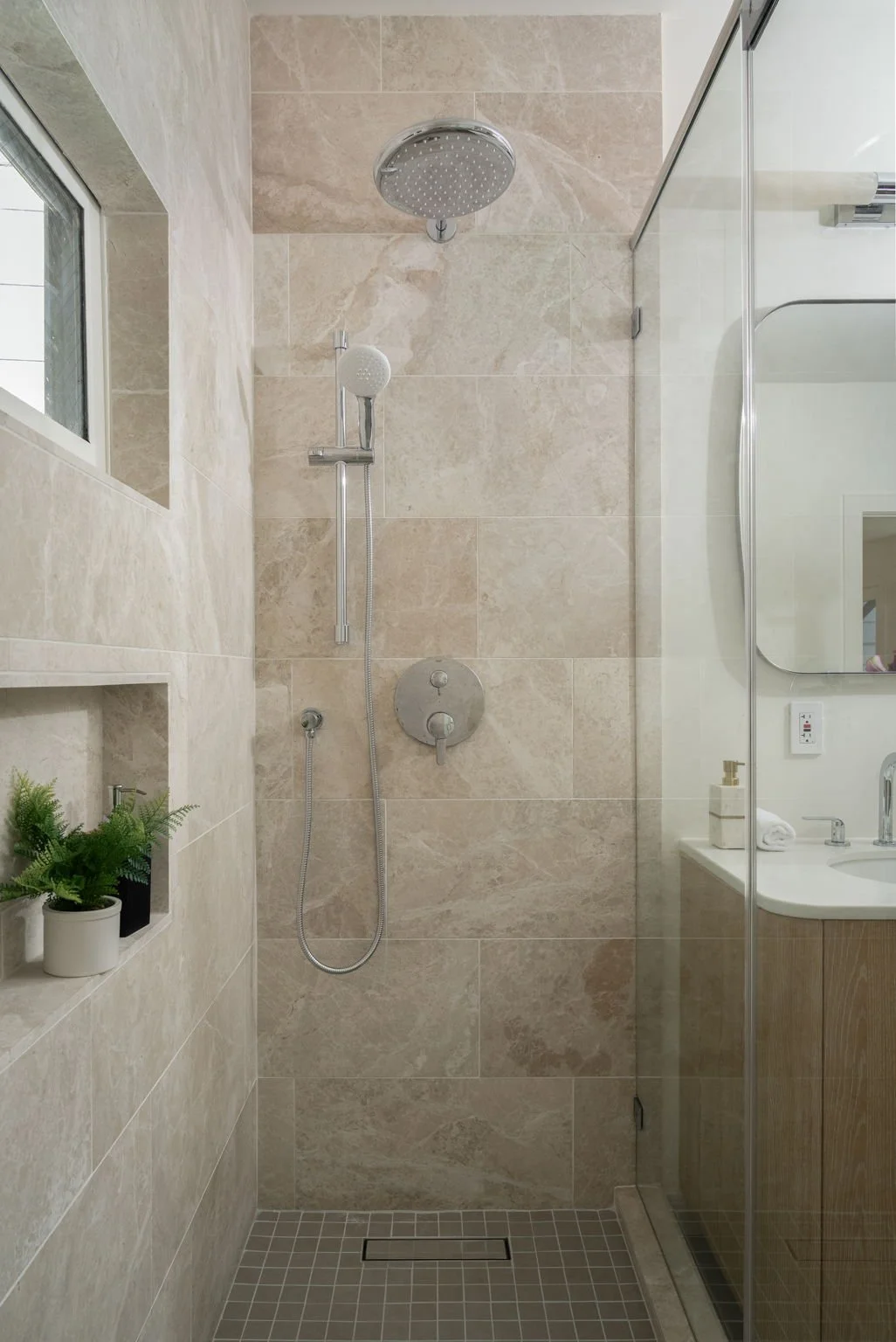Modern shower area with beige marble tiles, a round overhead shower, a handheld showerhead, a built-in wall shelf with small potted plants, adjacent to a bathroom sink and mirror, with a glass partition.