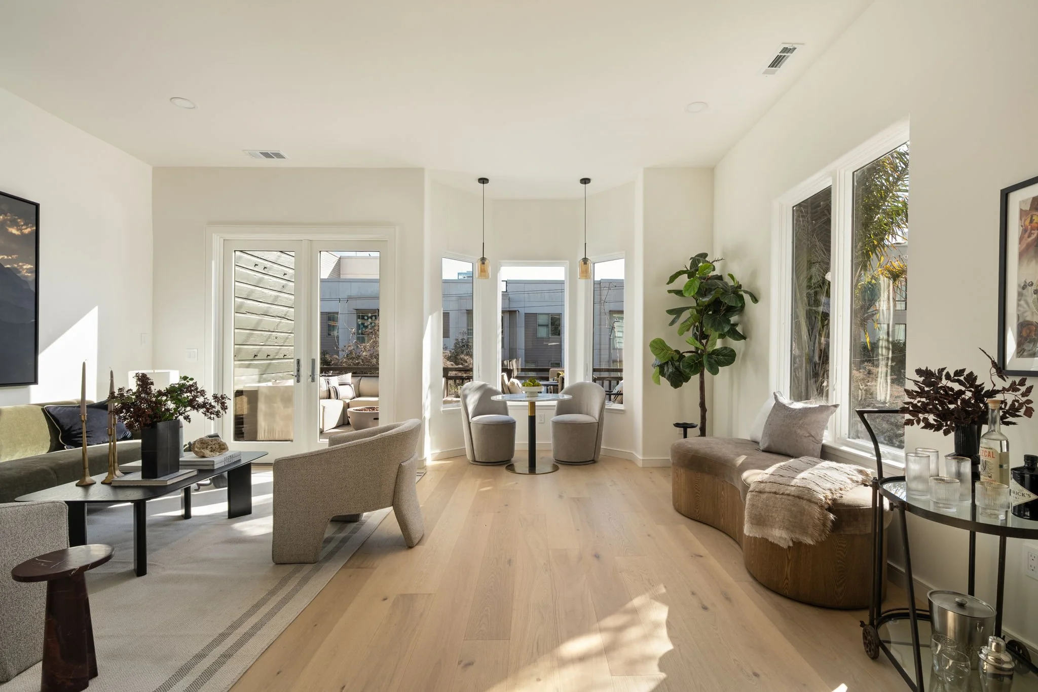 Bright living room with large windows, beige couches, a potted plant, and a dining area with a round table, all illuminated by natural sunlight.