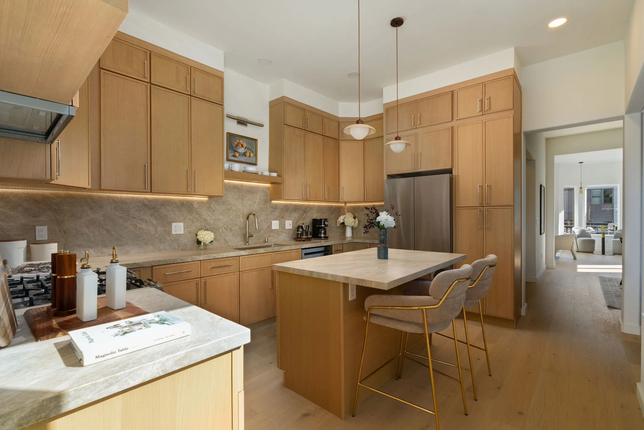 Modern kitchen with light wood cabinets, beige marble countertops, a central island with matching countertop, and three pendant lights hanging above. The kitchen includes a stainless steel refrigerator, farmhouse sink, and decor such as flowers and b
