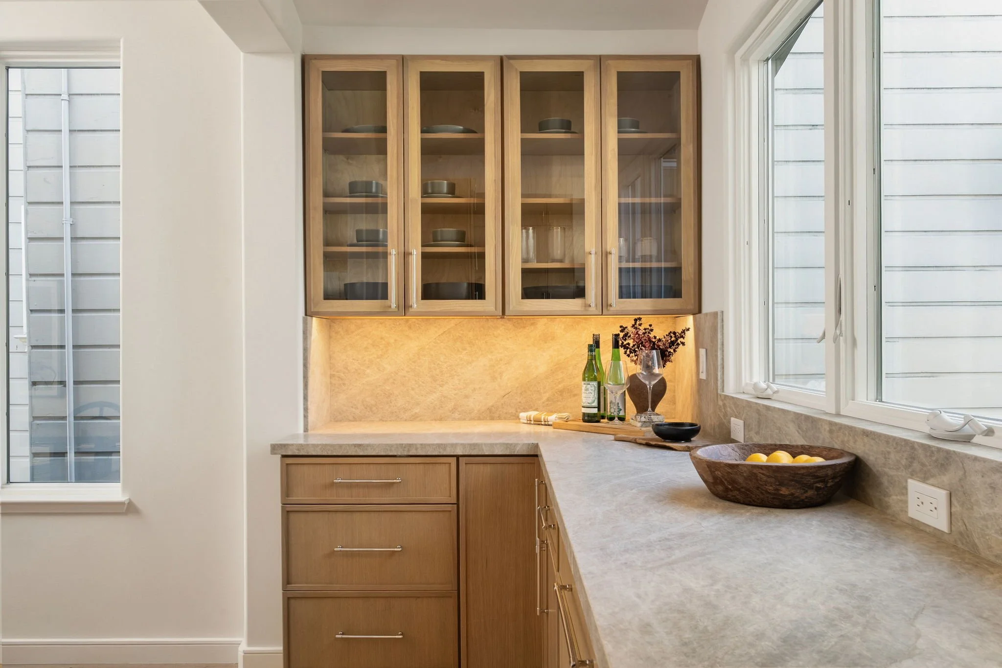 A kitchen with wooden cabinets, a book, a wine glass, and bottles on a marble countertop, near a window.