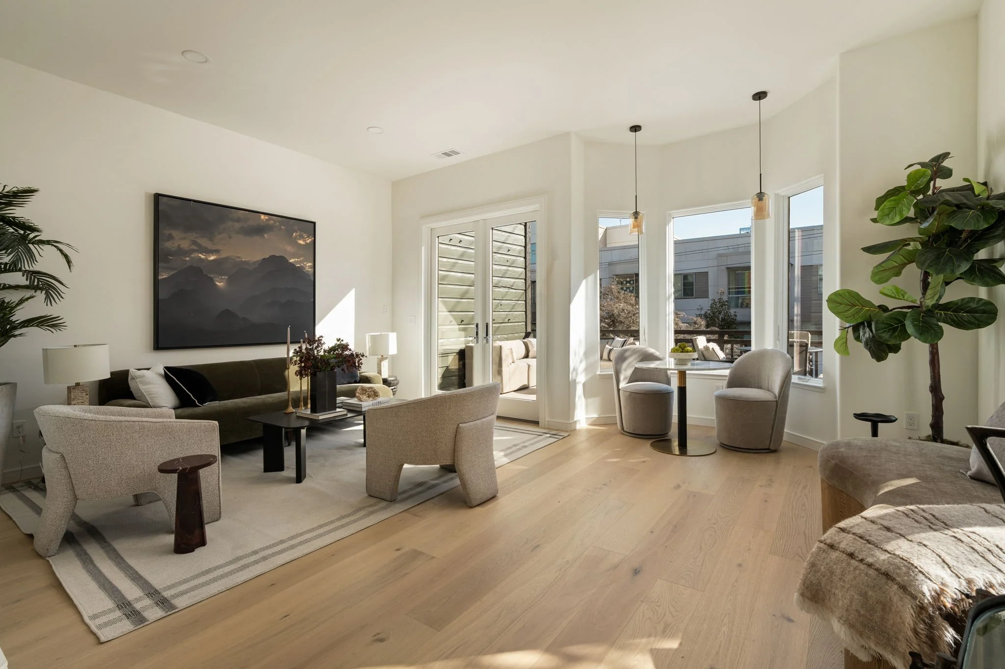 Bright living room with beige chairs, a black sofa, a large wall-mounted TV, and multiple windows letting in natural light, decorated with plants and artwork.