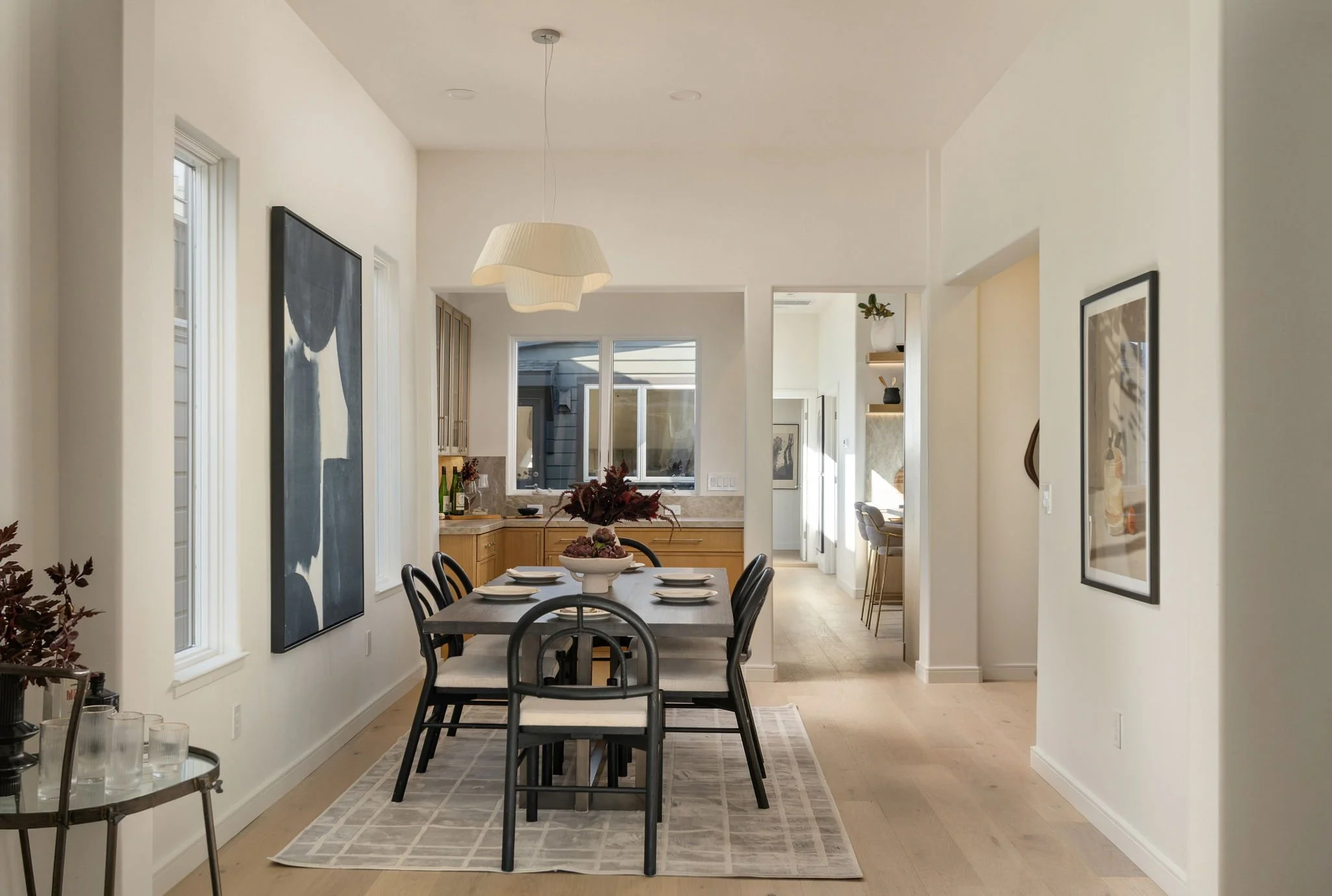 A dining area with a black dining table, six black chairs, and white plates. A decorative centerpiece with flowers is on the table. There is a radiator and a window in the background, and framed artwork on the walls. The room has light wood flooring, walls painted white, and a light-colored patterned rug beneath the table. A modern white pendant light hangs from the ceiling.