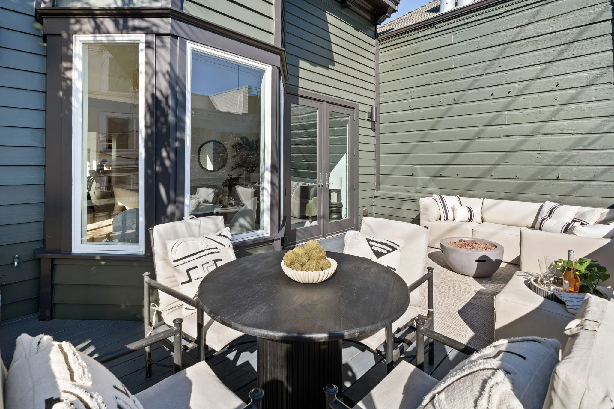 Outdoor patio area with beige sectional sofa, black round dining table with white cushions, potted plants, and glass of beverages, adjacent to a house with green wooden siding and large windows.