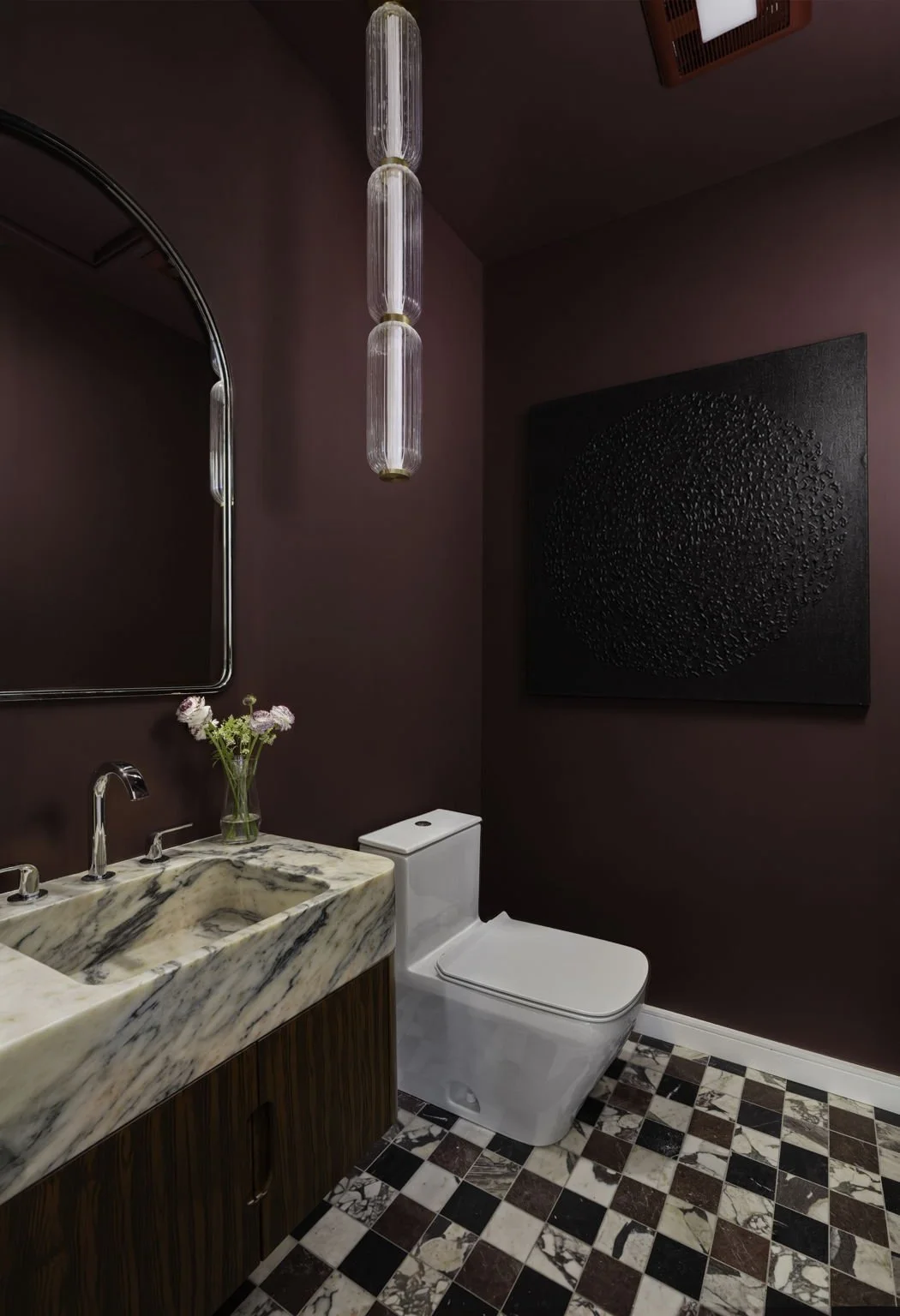 A stylish bathroom with burgundy walls, a marble sink, a modern white toilet, a black textured artwork, a mirror, a vase with flowers, and a hanging light fixture.