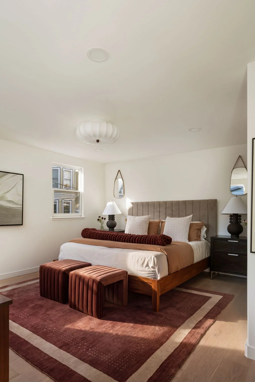 A modern bedroom with a large bed, two bedside tables with lamps, velvet footstools, a duvet, and decorative pillows. The room has a window, wall art, and mirrors on the wall, with a ceiling light fixture.