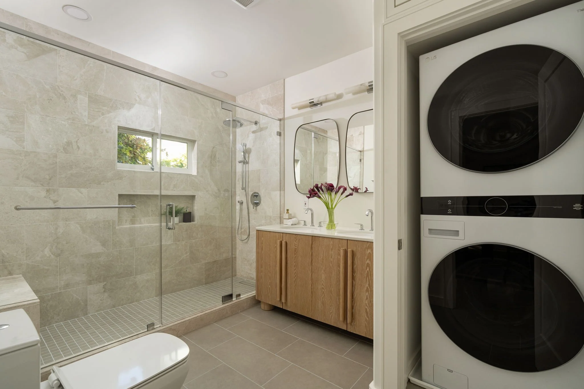 Bathroom with a glass-enclosed shower, vanity with a mirror and flowers, and stacked washer and dryer.