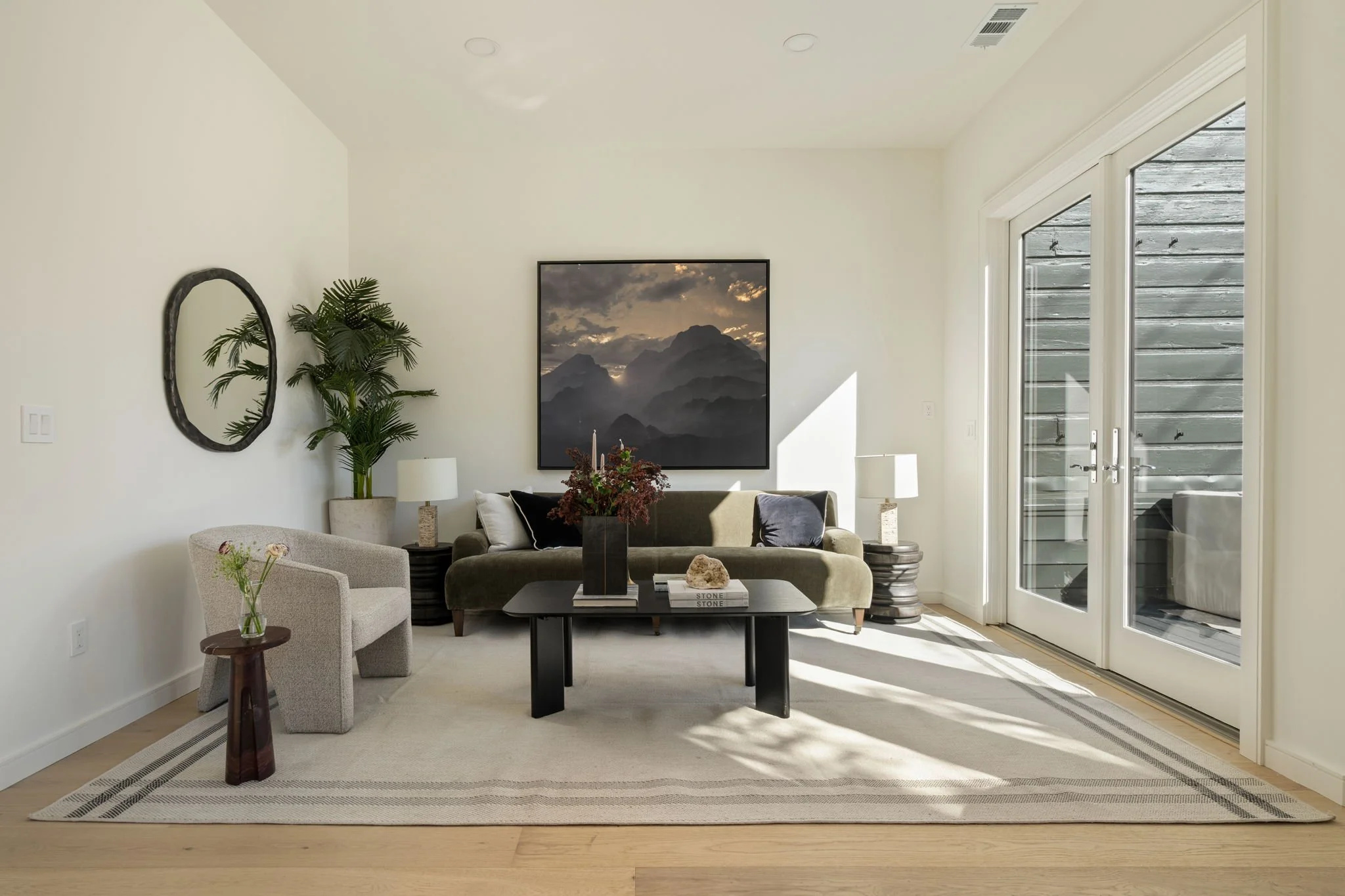 Living room with beige walls, a large window with sliding glass doors, a sofa, a coffee table with books and decorative items, two table lamps, a potted plant, wall art of mountains, and a round mirror.