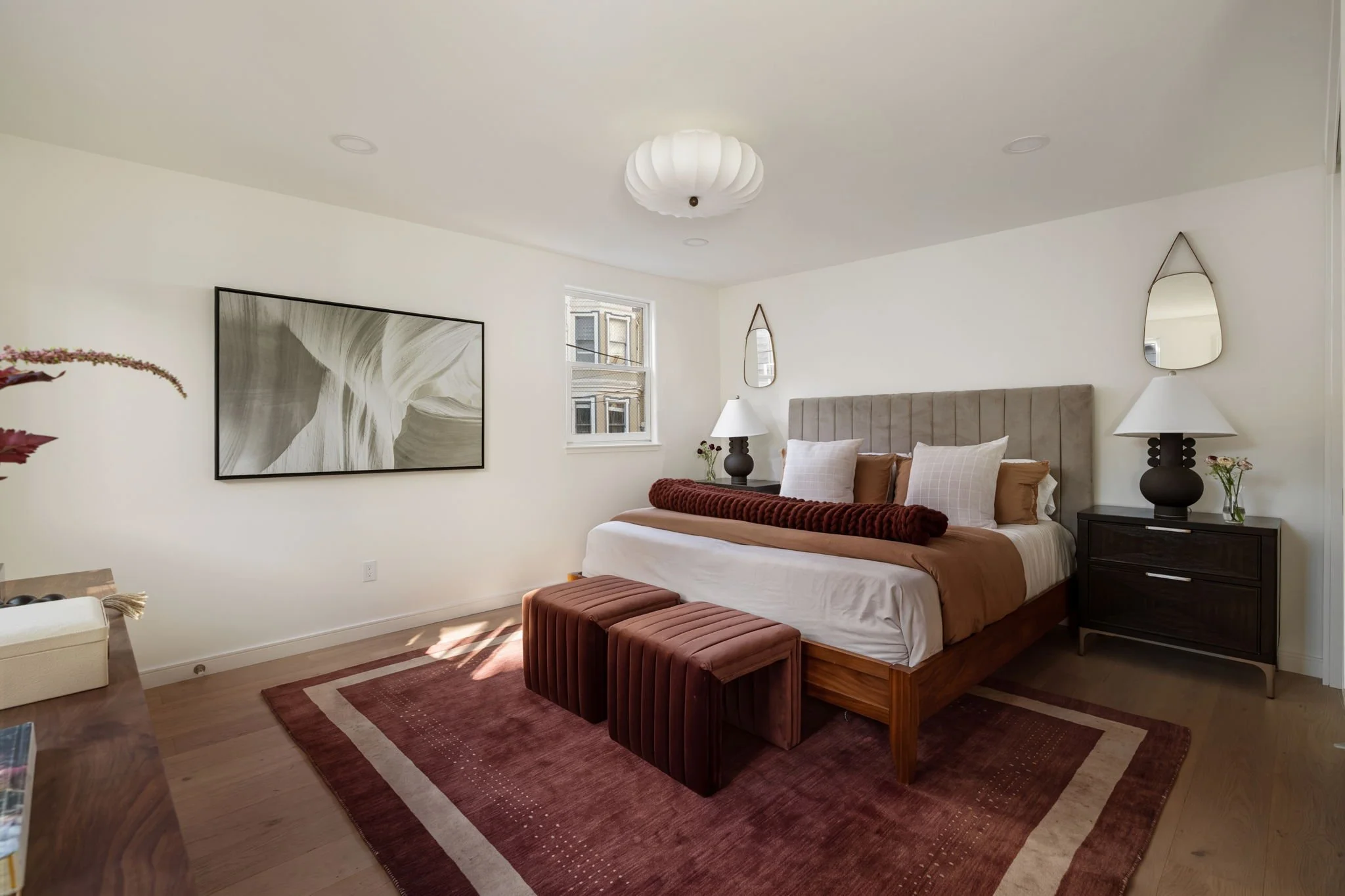 A bedroom with a bed, two nightstands with lamps, a wall mirror, a window, artwork, a velvet bench, a red area rug, and a white ceiling light fixture.