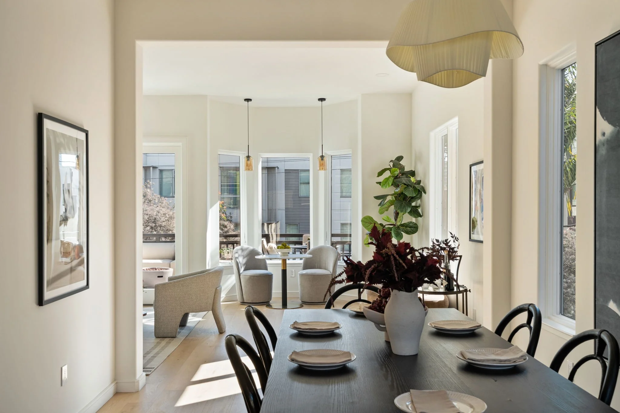 Interior view of a bright dining area with a table and chairs, modern light fixtures, potted plants, and large windows showing neighboring buildings.