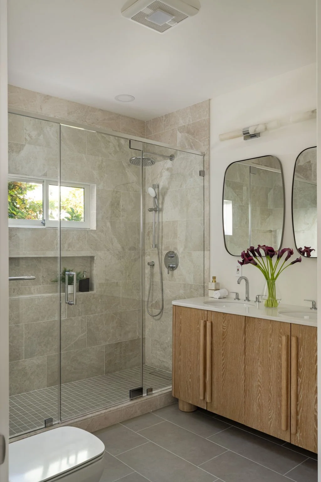 Modern bathroom with a glass shower enclosure, a wooden vanity with double sinks, two oval mirrors, and a vase of dark purple flowers on the counter.