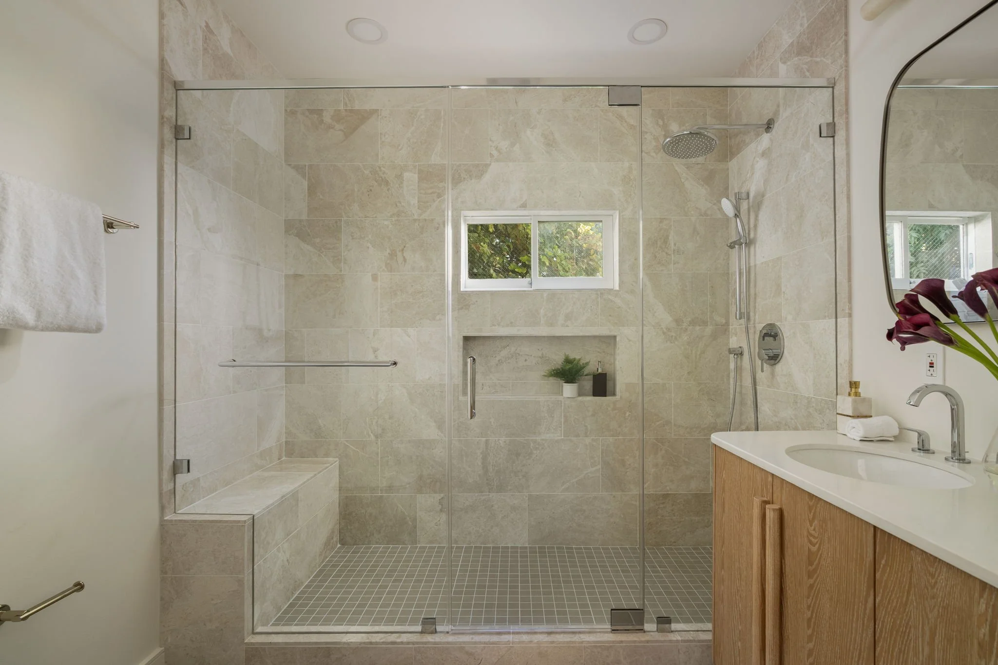 Modern bathroom with a walk-in shower enclosed by glass, beige tile walls, small square beige tile floor, a small window with greenery outside, a wooden vanity with a white sink, mirror, and a vase with flowers.