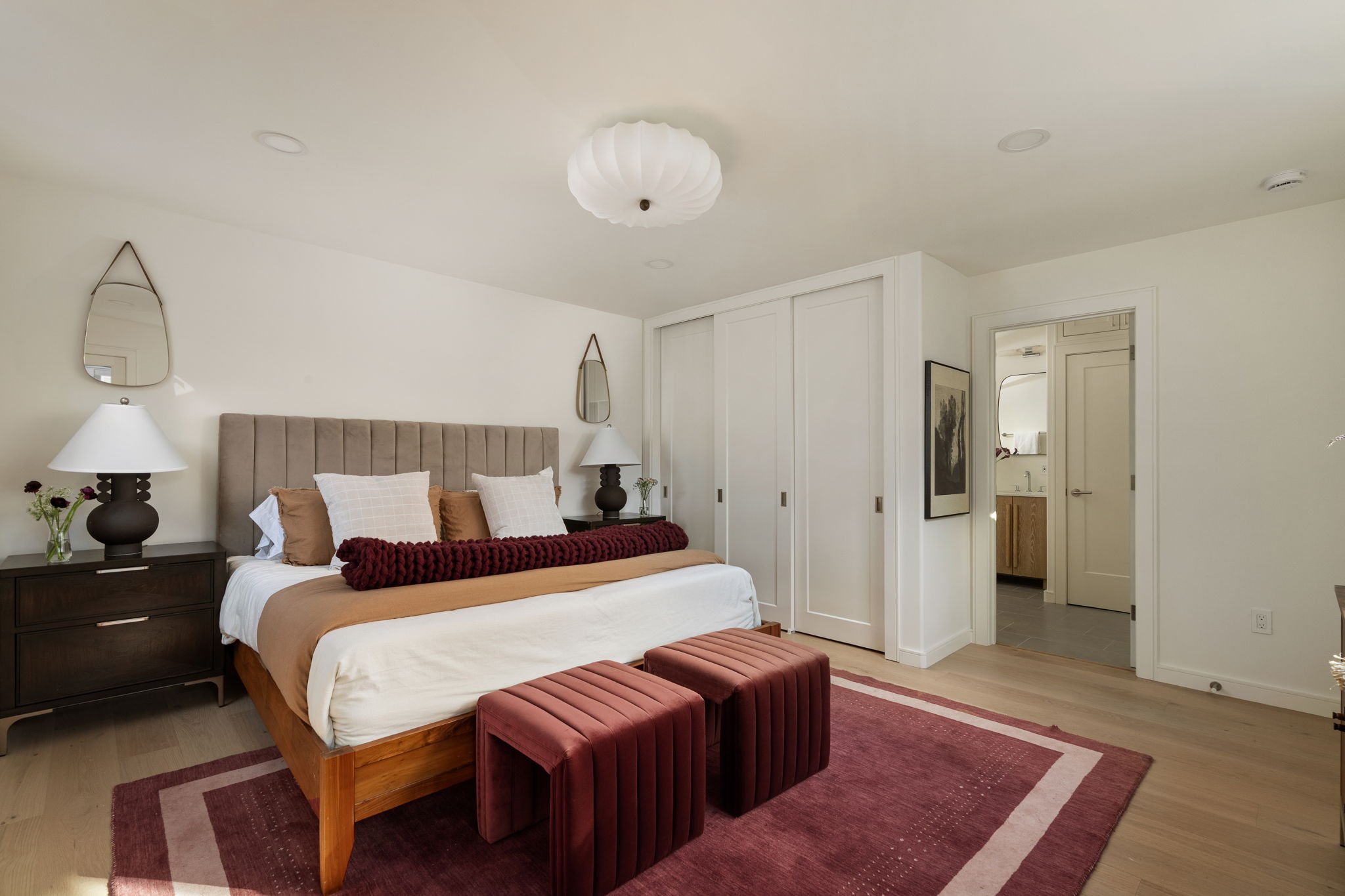 A bedroom with a bed featuring a tufted beige headboard, white bedding, and a tan blanket, flanked by matching black bedside tables with white lamps. Two reddish-brown upholstered benches are at the foot of the bed. The room has a red area rug, a large wall mirror, and a framed picture on the right wall. There are built-in white closets and an open doorway leading to a bathroom.