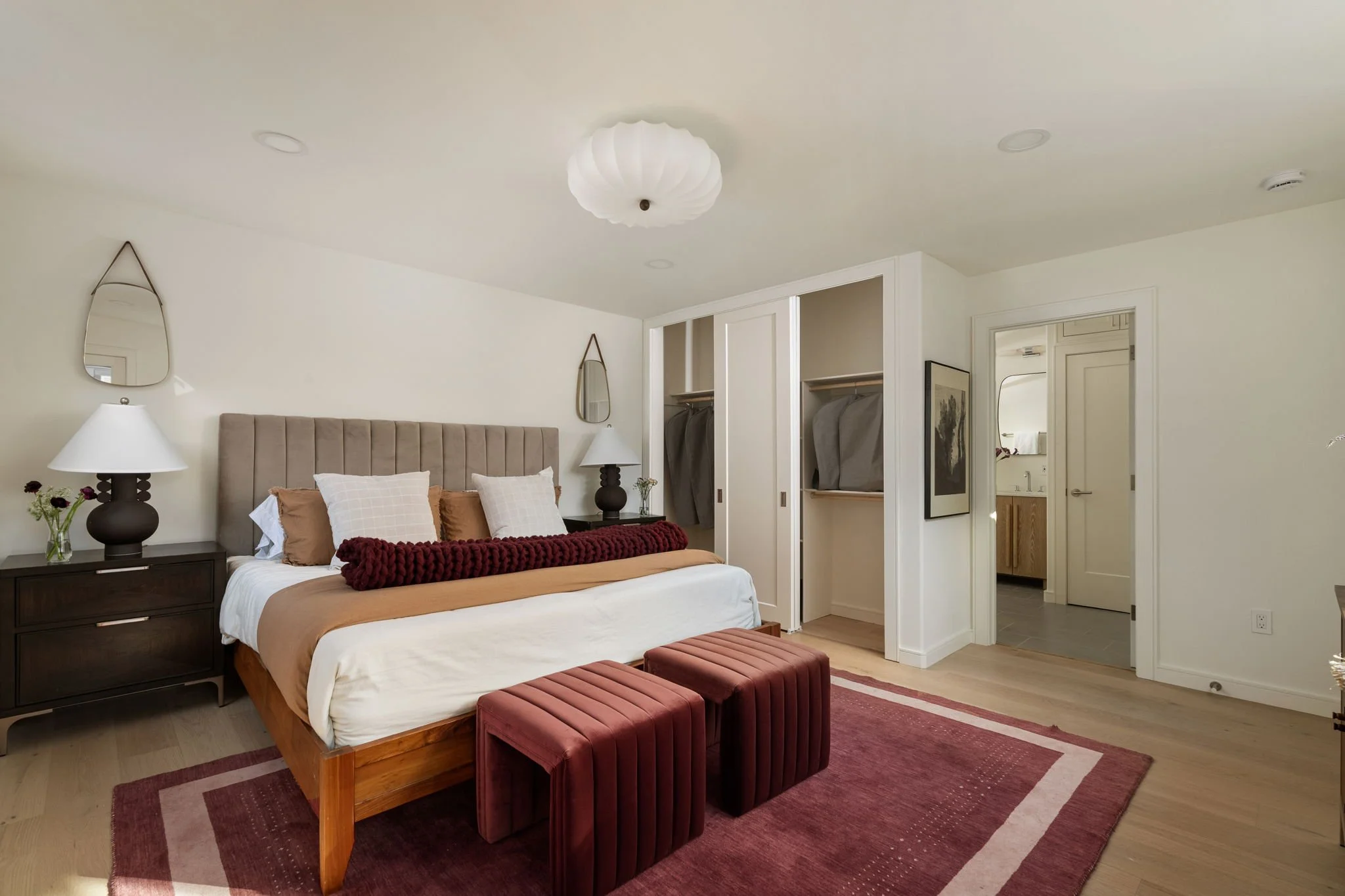 Modern bedroom with beige upholstered bed, dark wooden nightstands, white walls, a closet with sliding doors, and a doorway leading to a bathroom. The room has a red and beige area rug and red velvet benches at the foot of the bed.