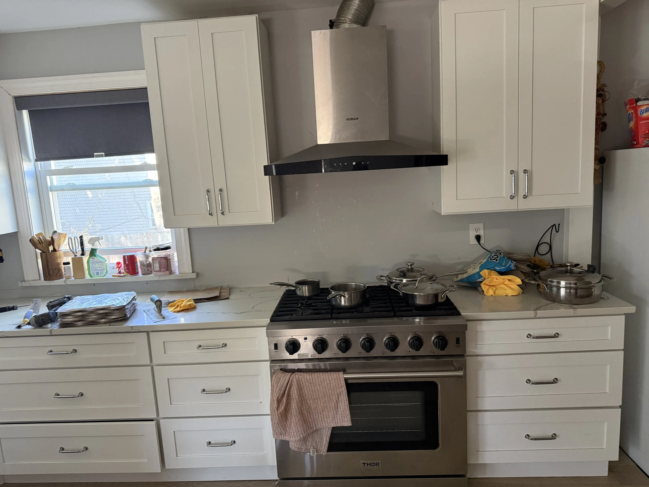 A kitchen with white cabinets, a gray marble countertop, and a stainless steel stove. Items such as pots, pans, utensils, and cleaning supplies are on the countertop. A window with a black shade is above the sink, and there are various items on the window sill, including bottles and containers.