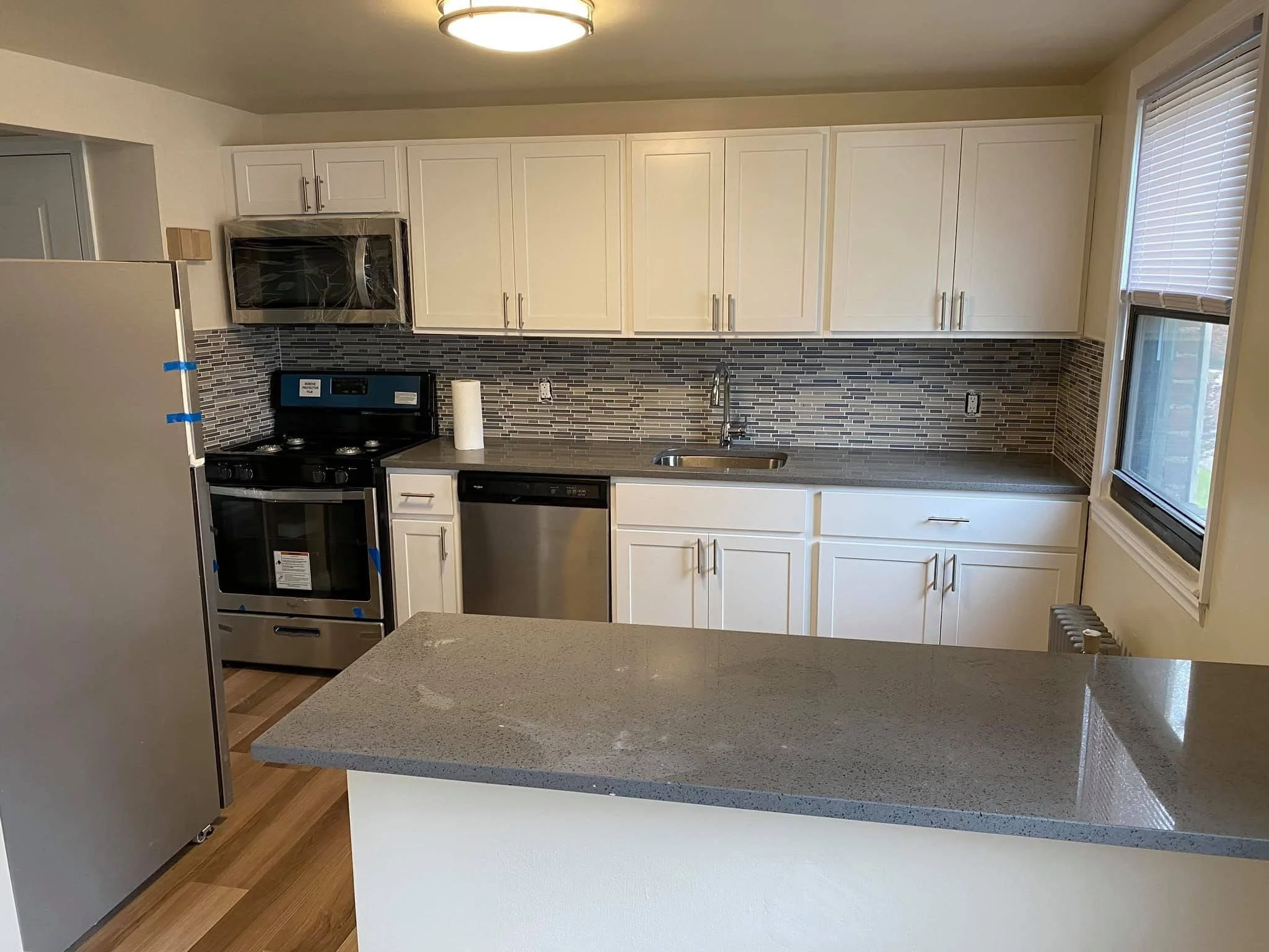 Modern kitchen with white cabinets, gray granite countertops, a gray backsplash, a stainless steel dishwasher, stove, microwave, and a window with blinds.