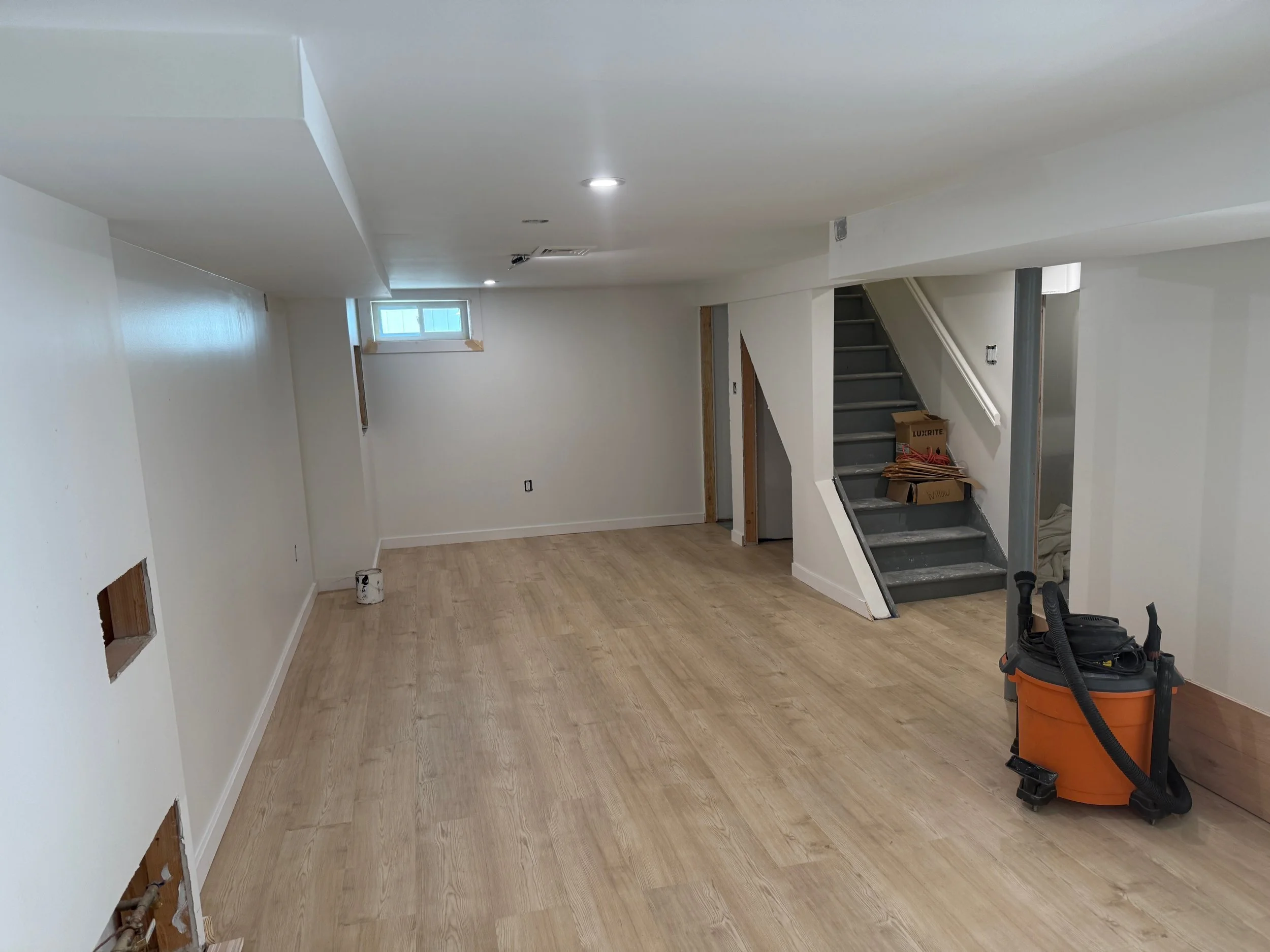 A room under renovation with unfinished walls, a staircase, and construction tools including a vacuum and a box of materials.