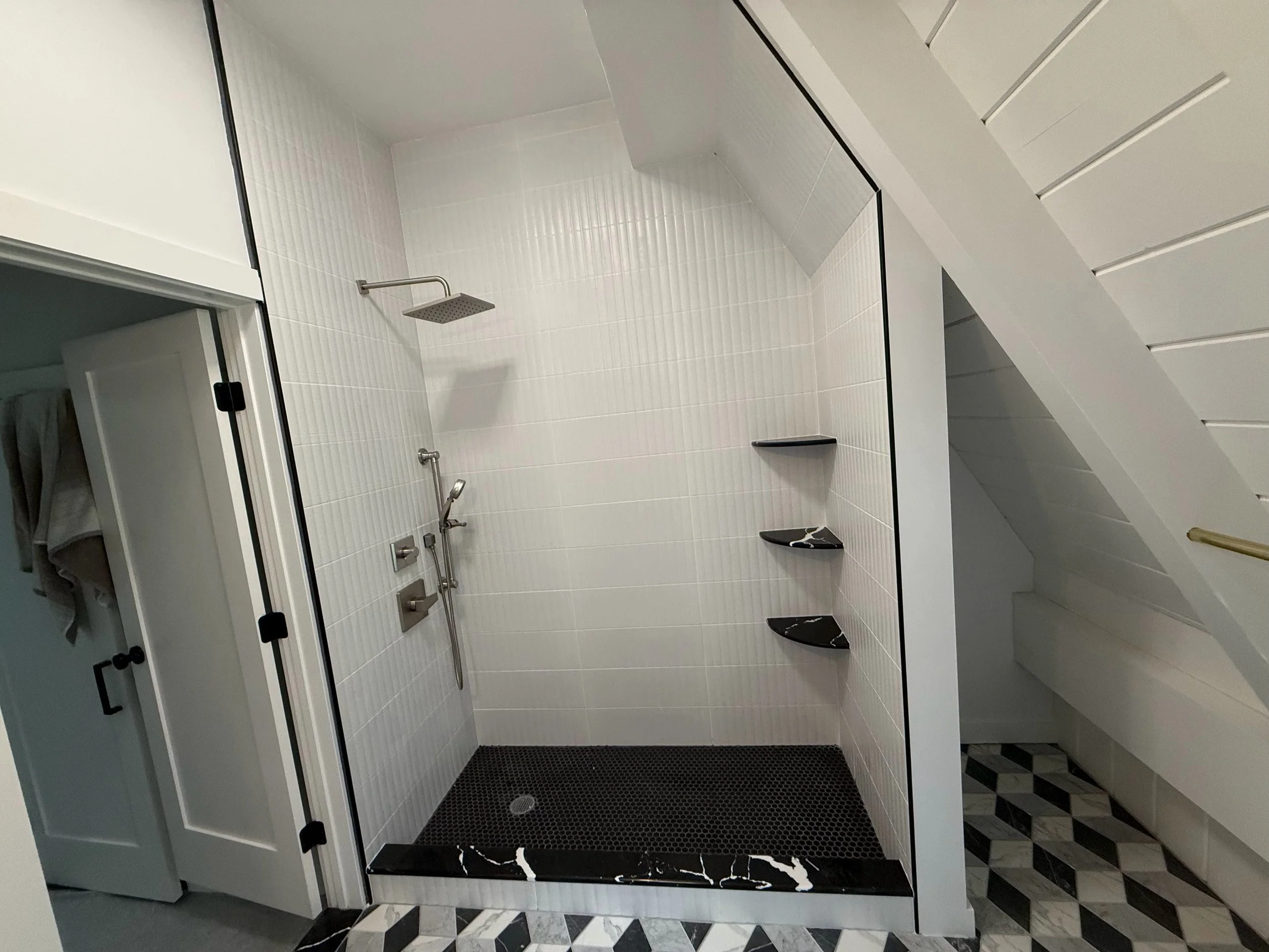 Modern shower area with white tiled walls, a black marble shelf, a rain showerhead, and a black rubber mat on the floor, adjacent to a toilet with black and white tile flooring.