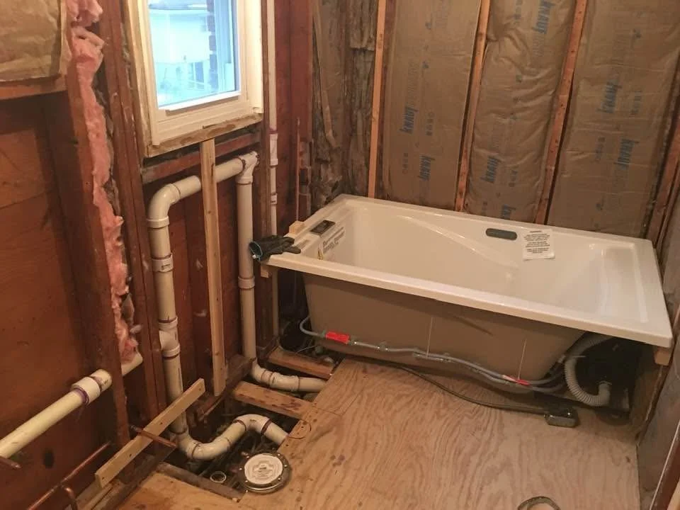 A bathroom with exposed wooden studs and insulation, featuring a white bathtub, plumbing pipes, and a small window.