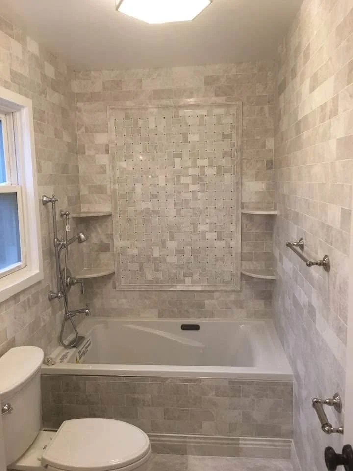 A small bathroom with a bathtub, a toilet, a window, and beige tile walls. The wall behind the bathtub has a decorative tile pattern and three corner shelves. There are metal towel bars on the walls.