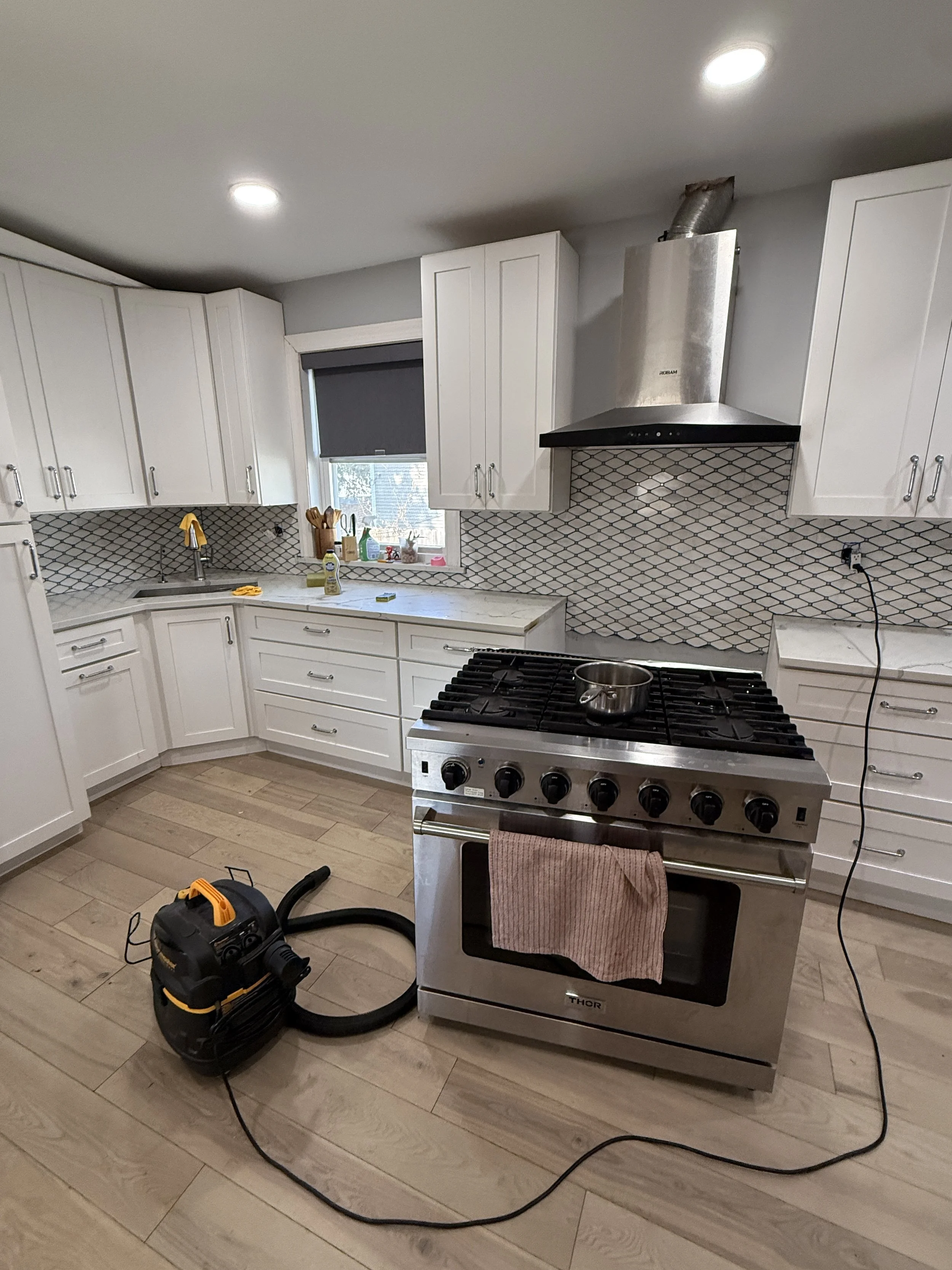 A kitchen with white cabinets, a black and white diamond tile backsplash, a window with gray blinds, and a stainless steel stove with a towel hanging from its handle. A vacuum cleaner is on the floor.
