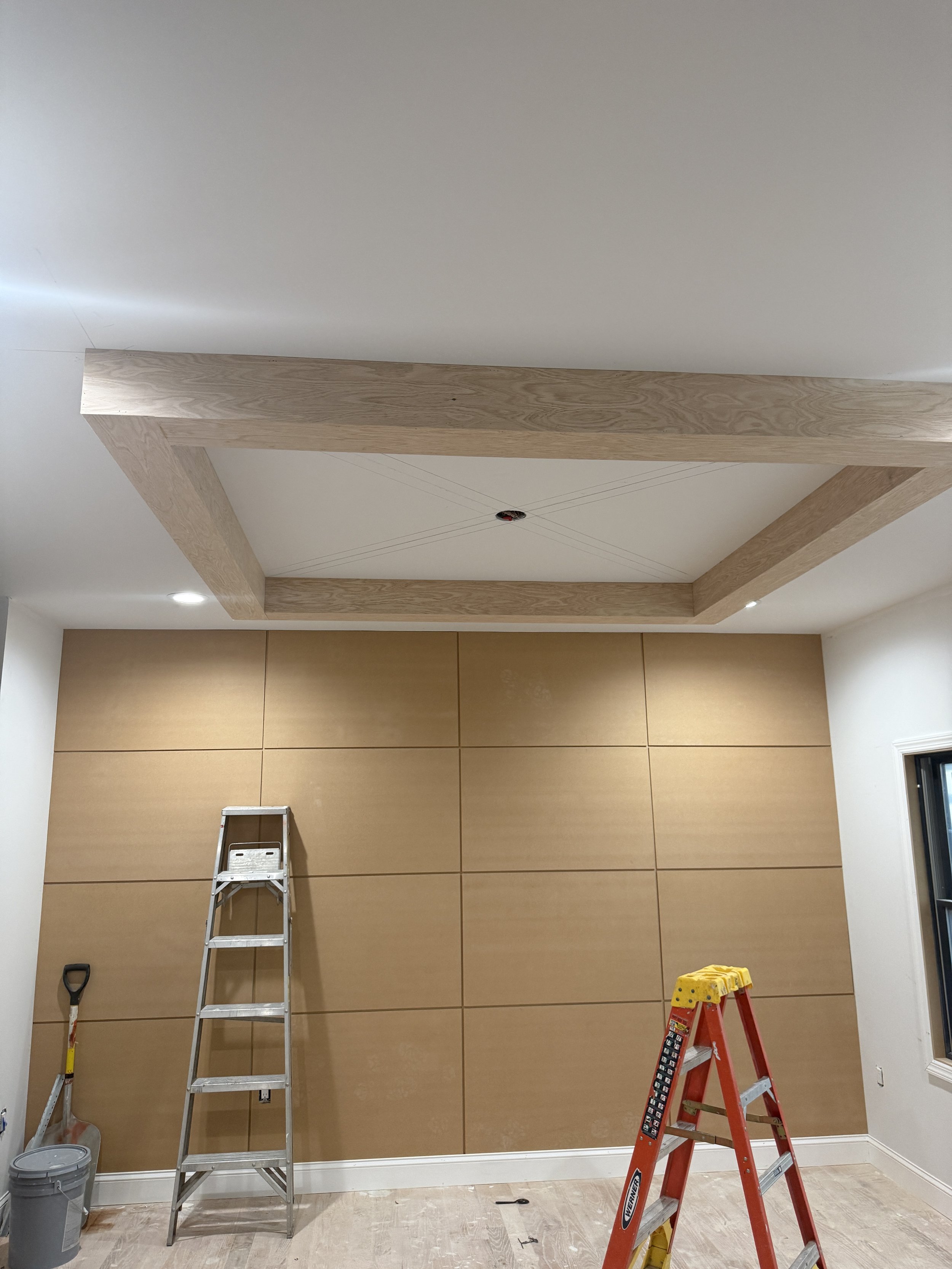 Interior of a room under renovation with beige wall panels, a stepped wooden ceiling frame, a ladder, and a red stepladder with a yellow toolbox on top.