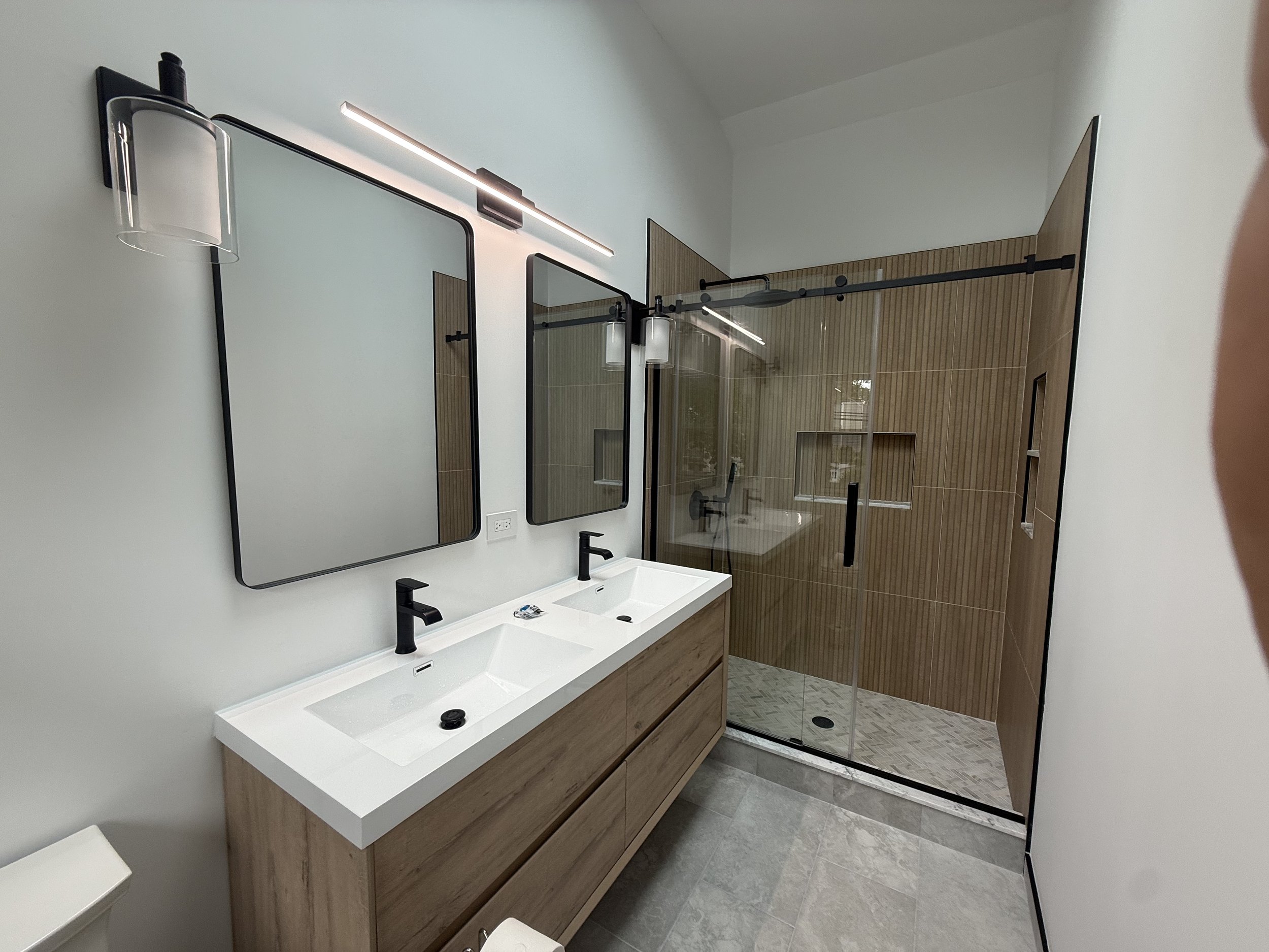 Modern bathroom with a white rectangular sink, black faucet, mirror, wall-mounted light, wooden vanity, glass shower enclosure, wood-paneled wall, and tiled floor.