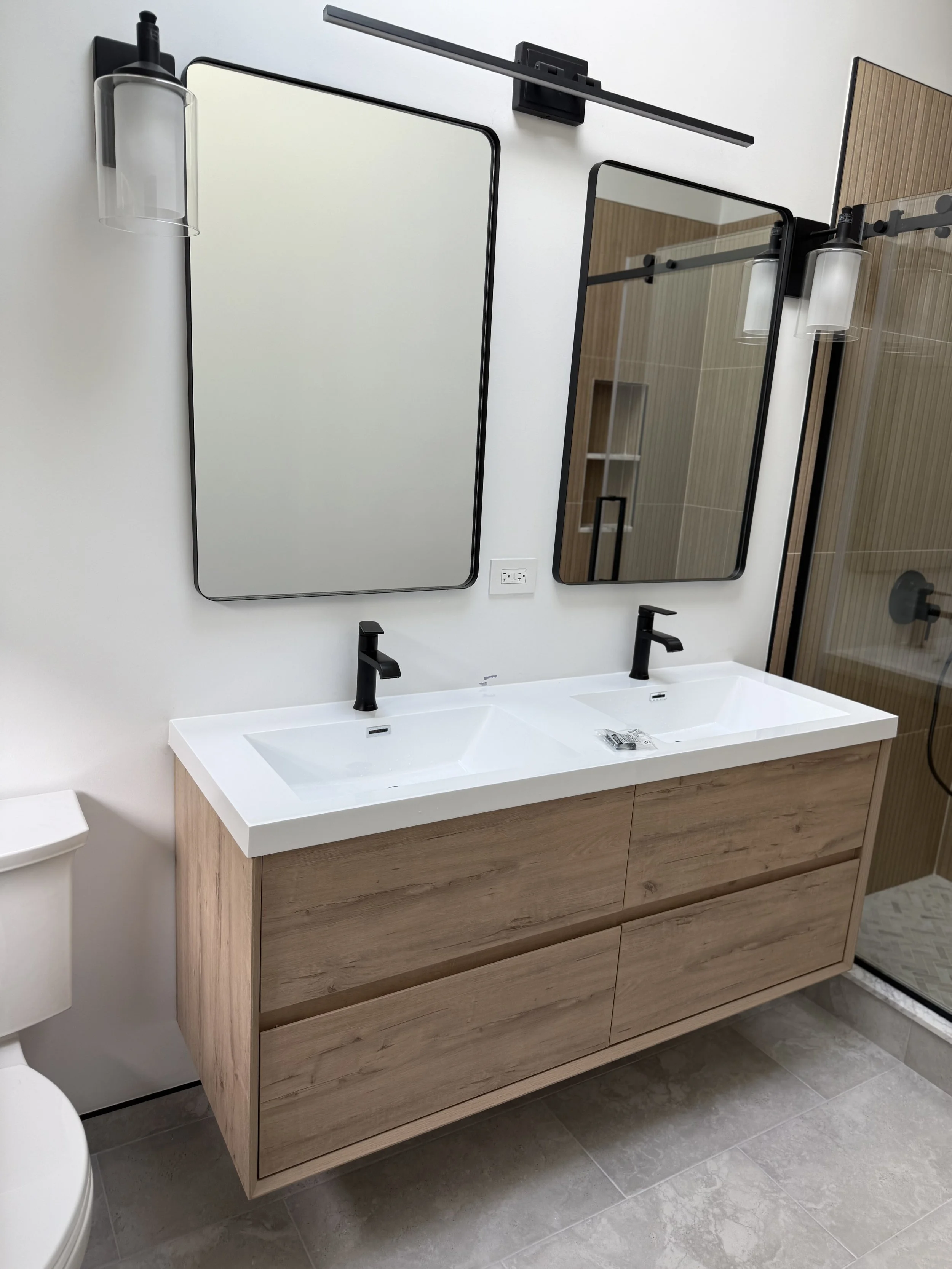 A modern bathroom vanity with a double sink, black faucets, a wooden cabinet, and two large black-framed mirrors mounted above the sinks. There are two black light fixtures with clear glass shades on either side of the mirrors. The wall behind is white, and the floor is gray tiled.