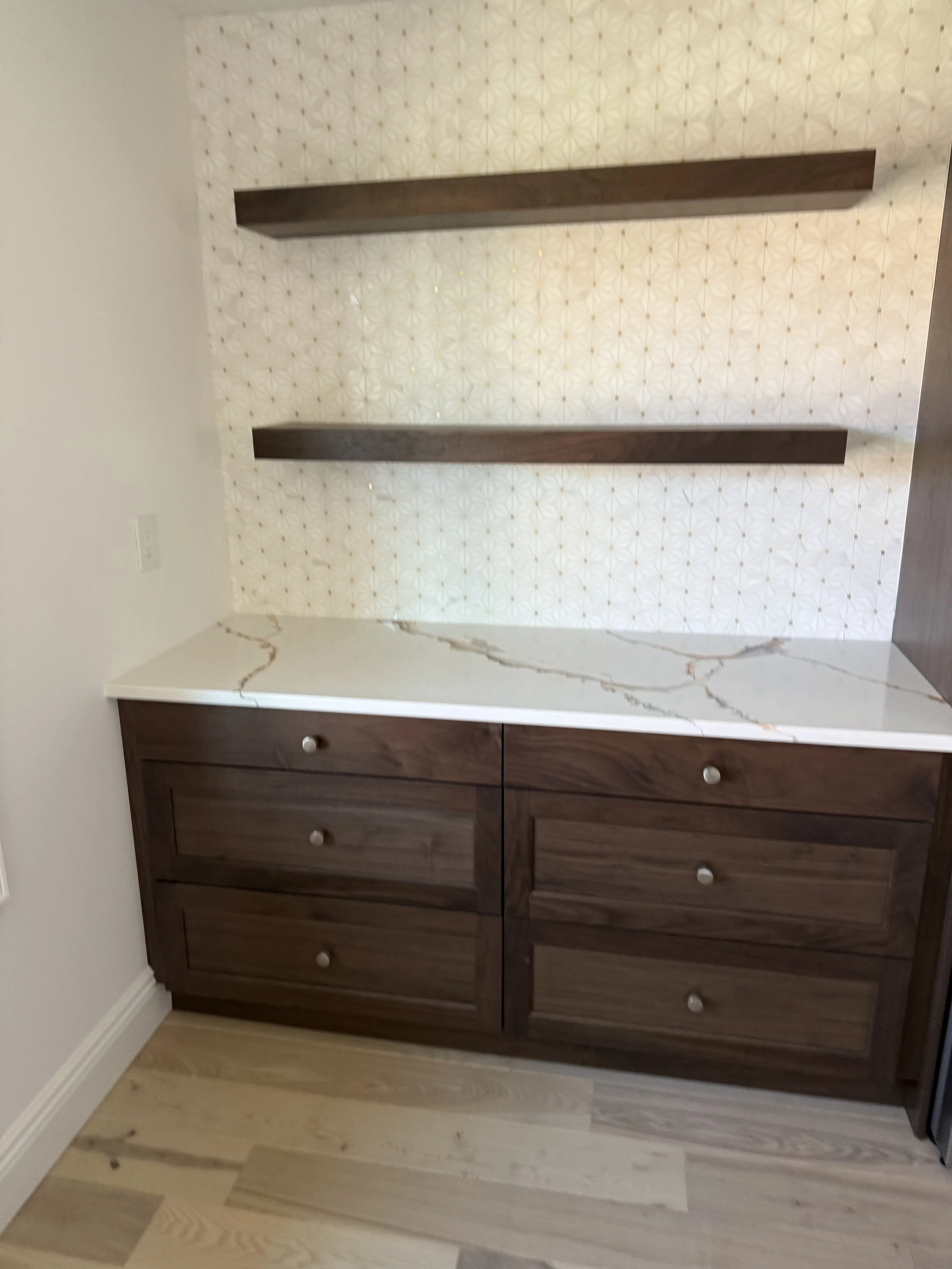 A dark wooden dresser with six drawers and silver knobs, placed against a wall with patterned wallpaper and a marble-patterned counter on top, with a light wood floor beneath.
