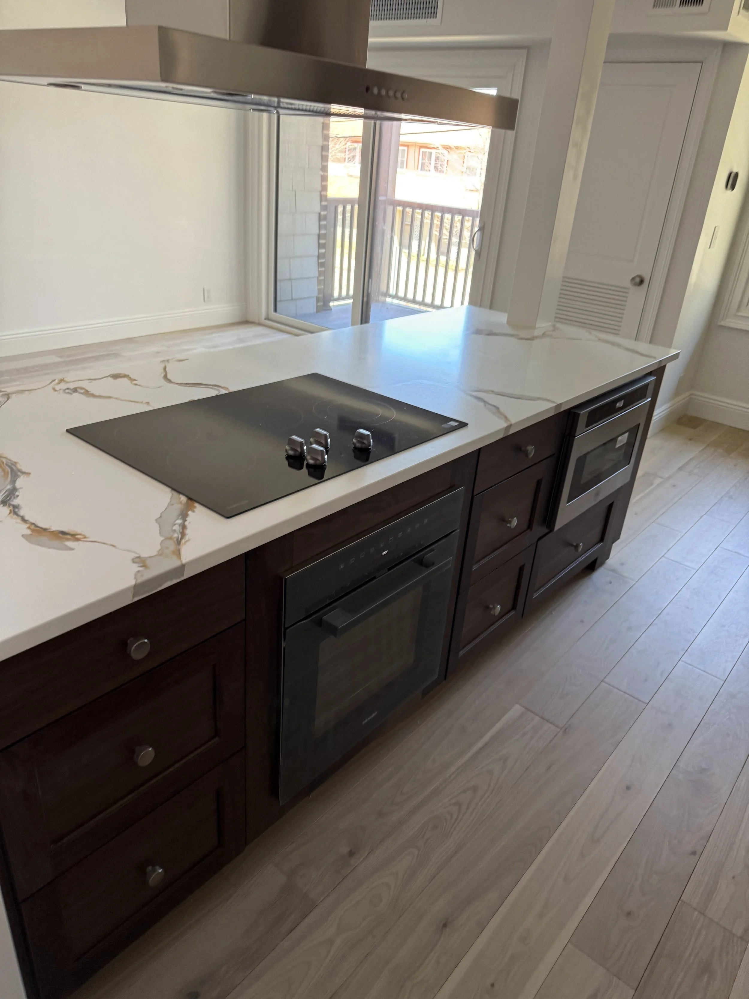A kitchen with dark wood cabinets, a white marble countertop with gold veining, a black electric cooktop, a built-in oven, a window overlooking a balcony with a railing, and light-colored wood flooring.