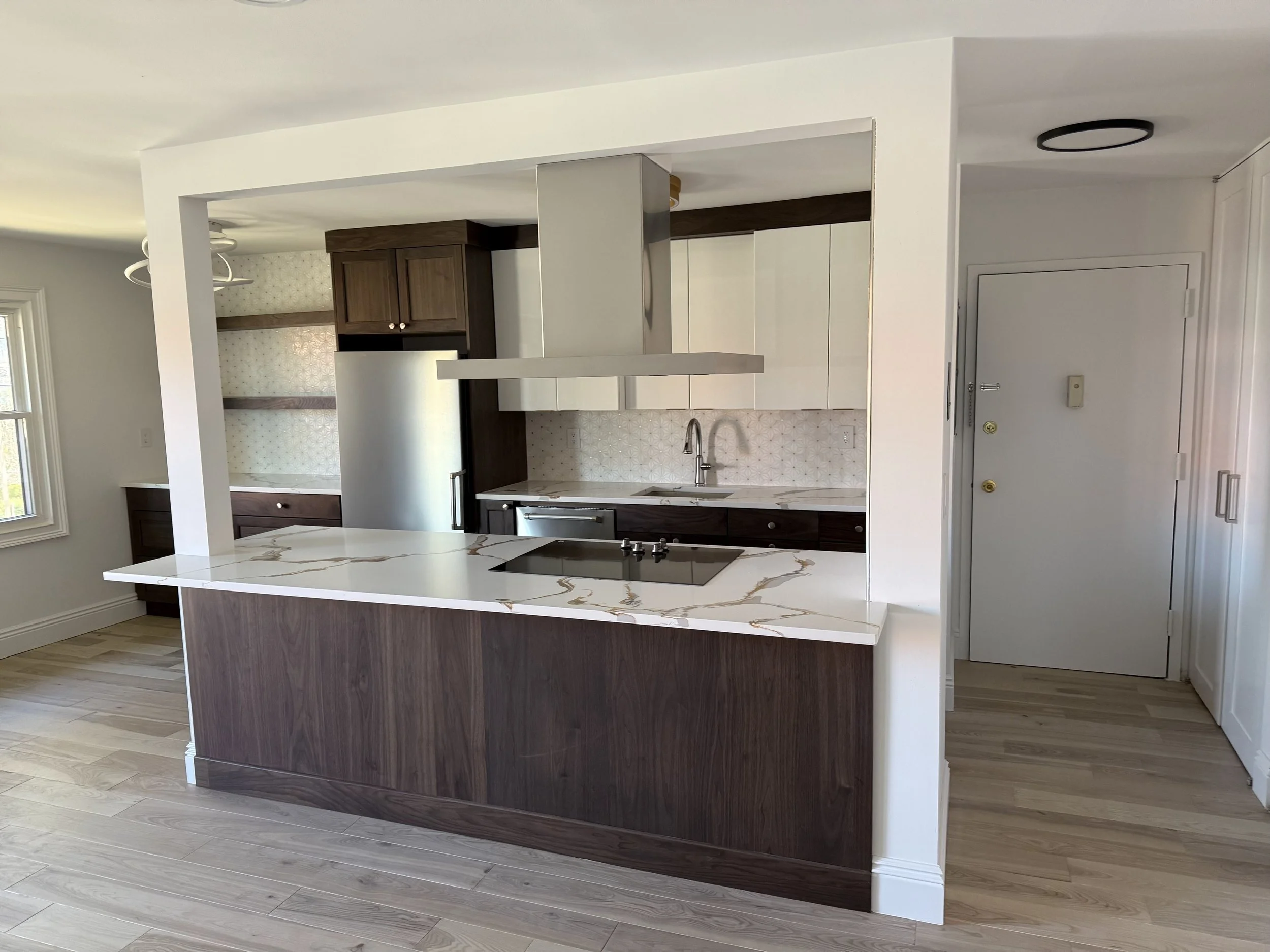 A modern kitchen with a white marble island, dark wood cabinets, and a white tiled backsplash with a geometric pattern. There's a window on the left and a closed door on the right.