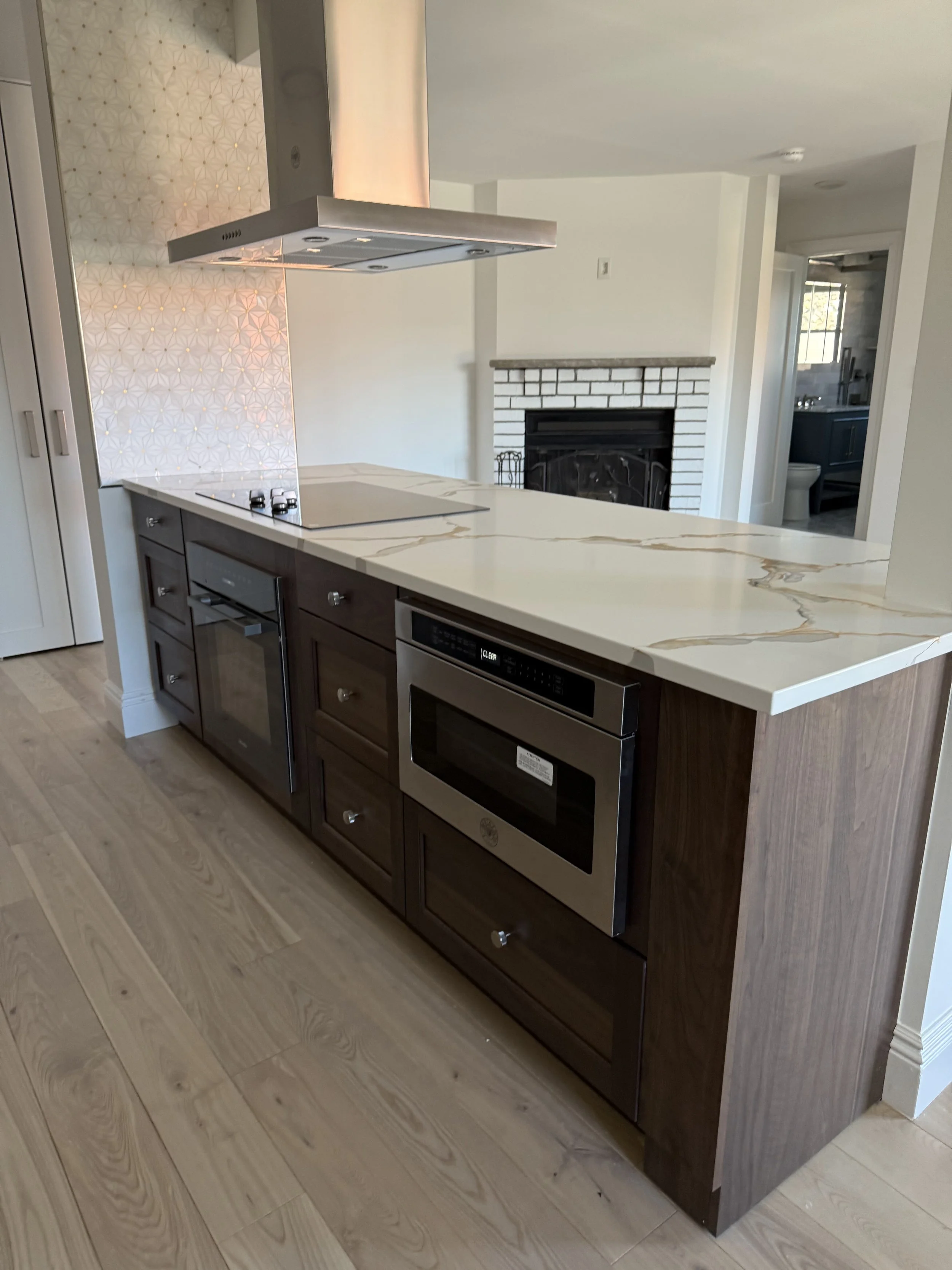 Modern kitchen island with a white marble countertop, dark wood cabinets, a built-in oven, and a cooktop with a stainless steel range hood above. In the background, there is a white brick fireplace and a doorway leading to a bathroom.
